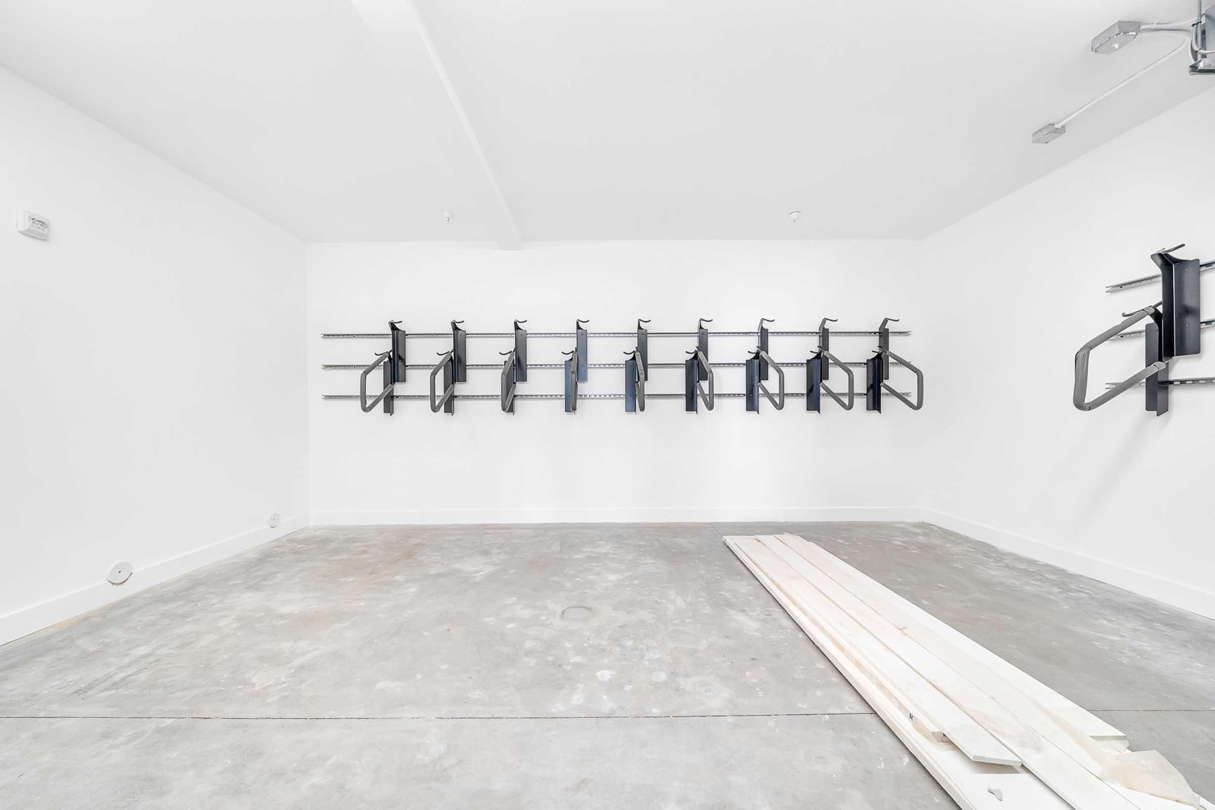 Empty garage with white walls, concrete floor, and black wall-mounted bike racks. A few long wooden planks are lying on the floor, and the space is well-lit, creating a clean and organized appearance.
