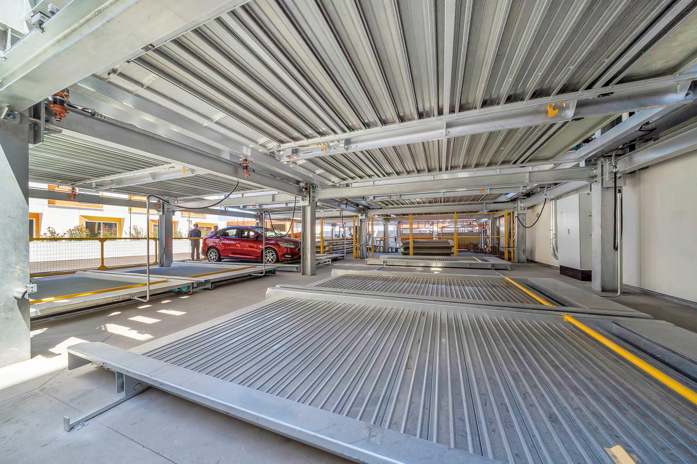 A multi-level parking structure with empty parking spaces and one red car parked on a lower level. The space features metal frameworks and ramps, illuminated by natural light. The design is modern and efficient, maximizing parking capacity.