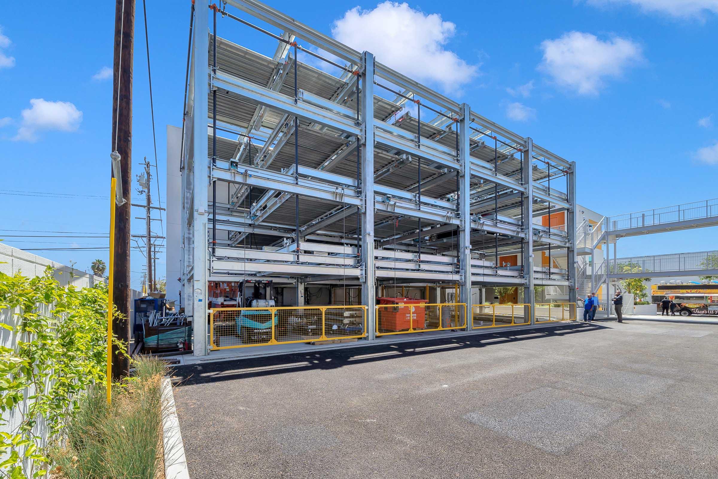 A modern multi-level parking structure with a steel frame and open design, surrounded by a paved lot. The structure features a combination of metal and glass elements and is set against a clear blue sky, with a person in blue clothing walking nearby.