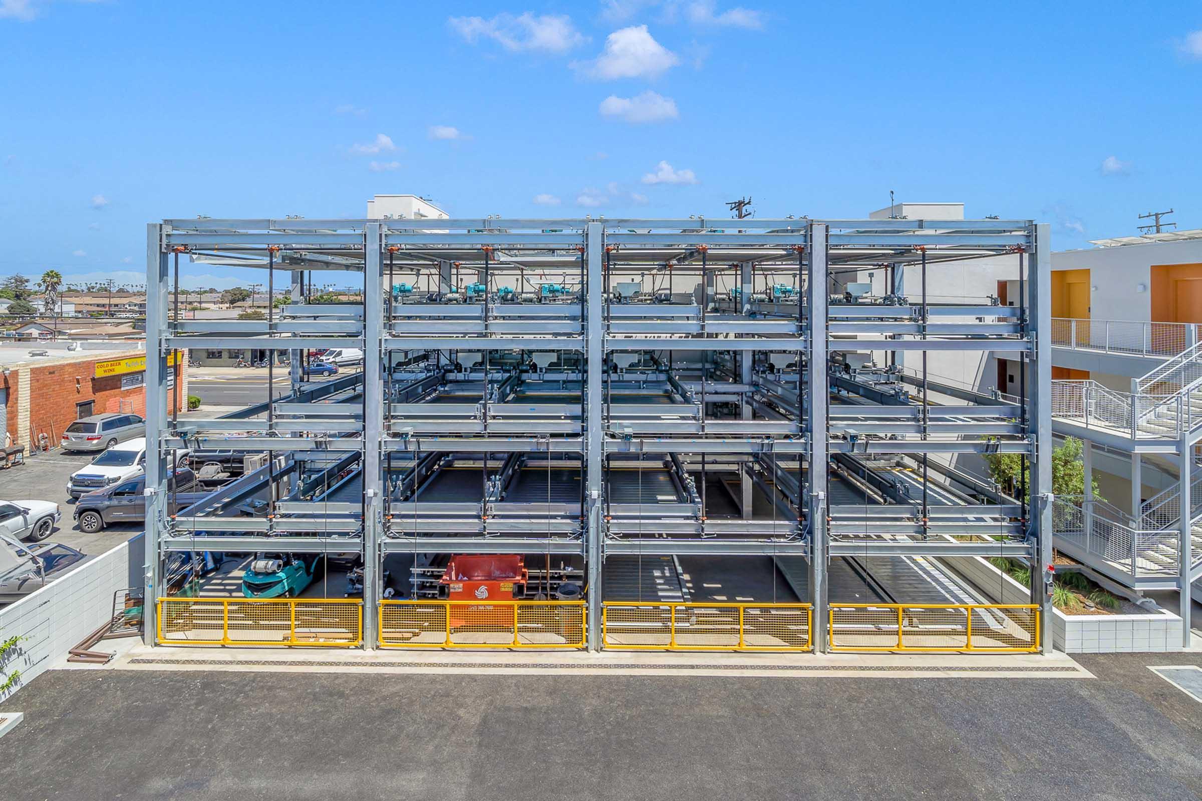 A multi-story automated parking structure with multiple levels of parking slots. The building has a modern design with a steel frame and is situated in an urban area. Clear blue sky above, with a few clouds visible. Nearby buildings and a parking lot can be seen in the background.