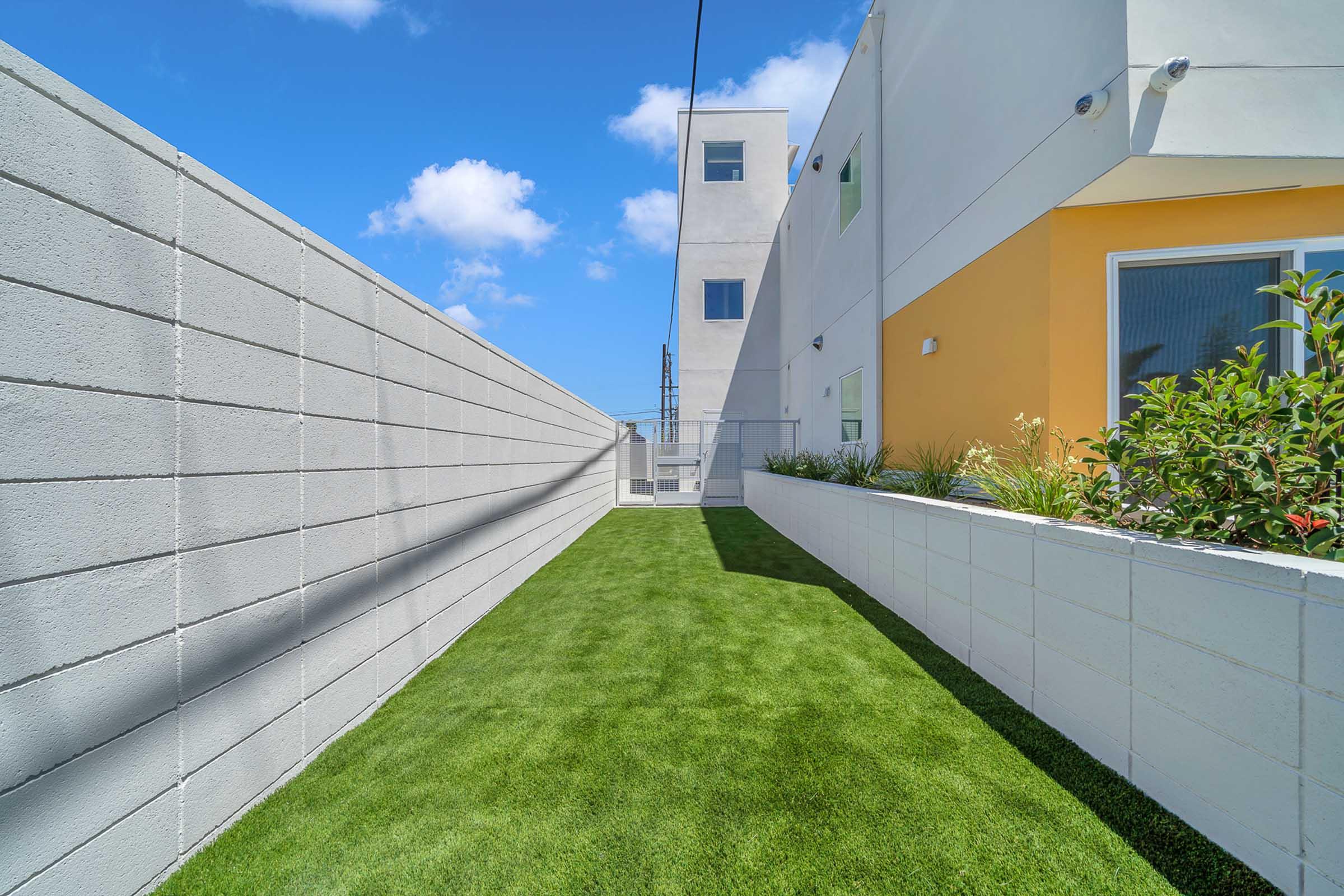 Narrow pathway lined with a white wall on one side and a modern building with a yellow accent on the other. The ground features lush green artificial grass, and bright blue sky with fluffy white clouds is in the background.
