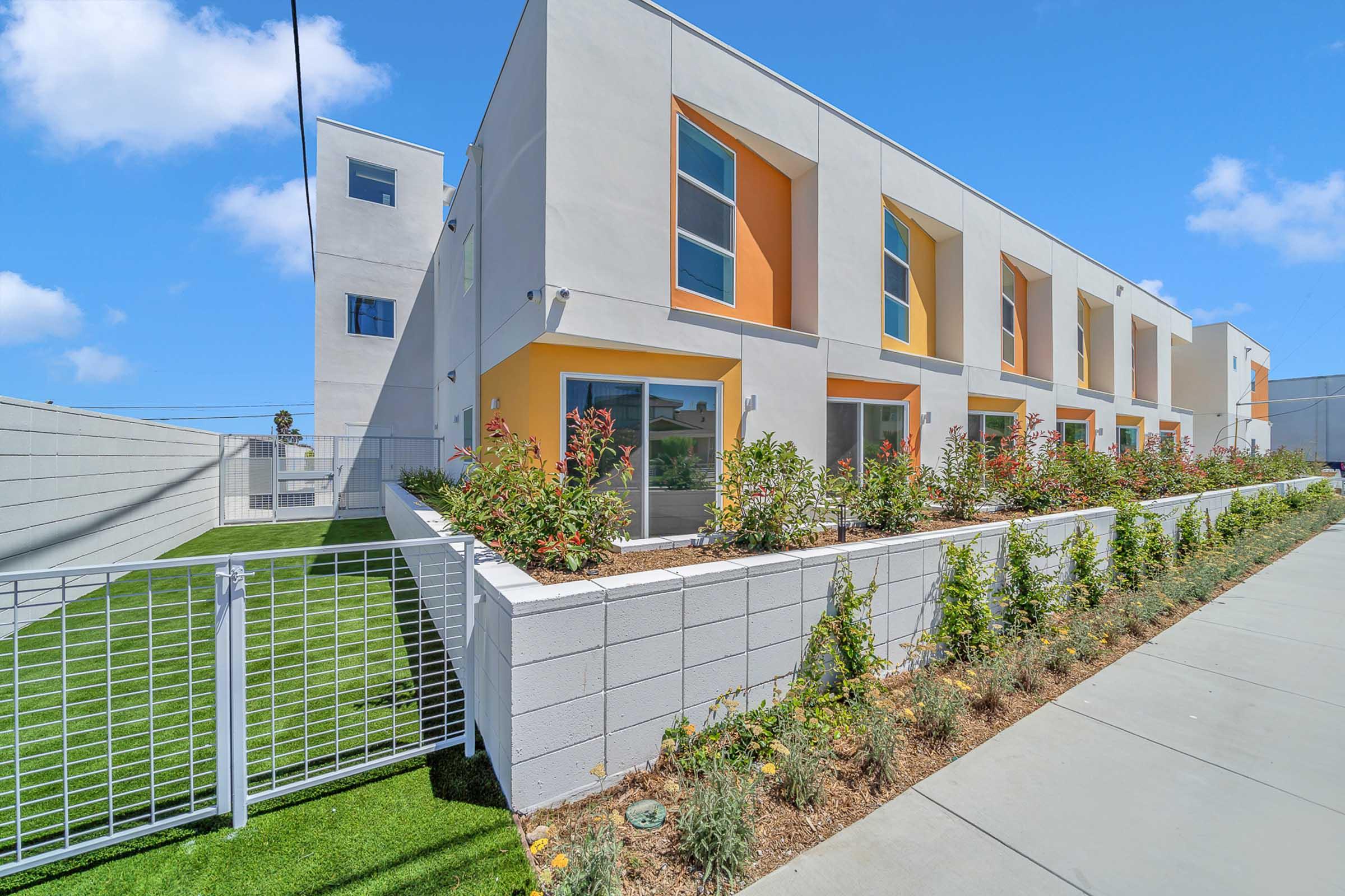 Modern residential building with a clean design featuring multiple windows and bright orange accents. Surrounding the building are landscaped areas with greenery and flower beds, along with a white fence. The sky is clear with a few clouds, creating a bright and inviting atmosphere.