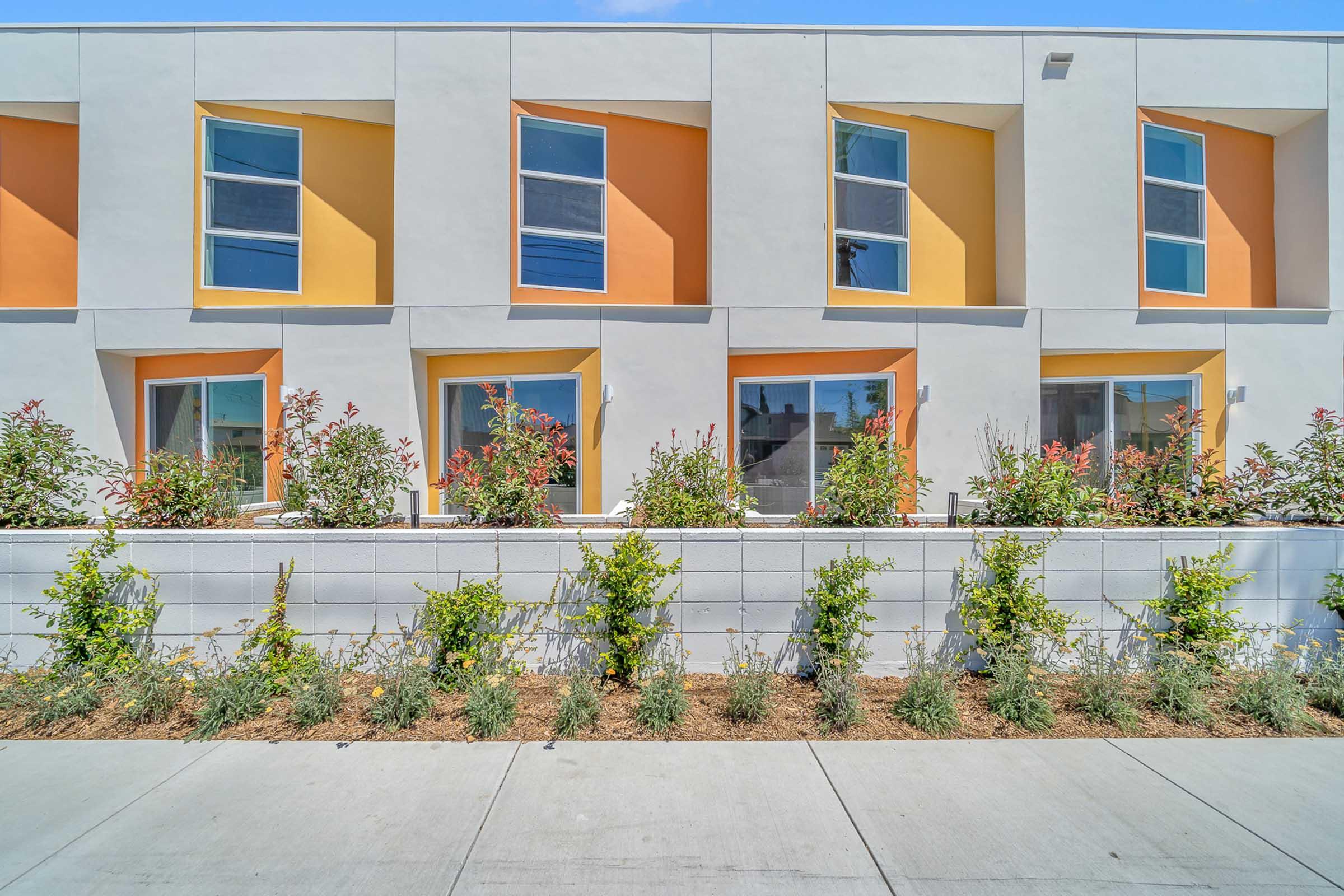 A modern residential building with a white and orange facade. The structure features multiple windows, some with angular designs. A neatly landscaped area in front includes low shrubbery and flowers, separated from the sidewalk by a low, gray wall. Bright blue sky above creates a vibrant atmosphere.