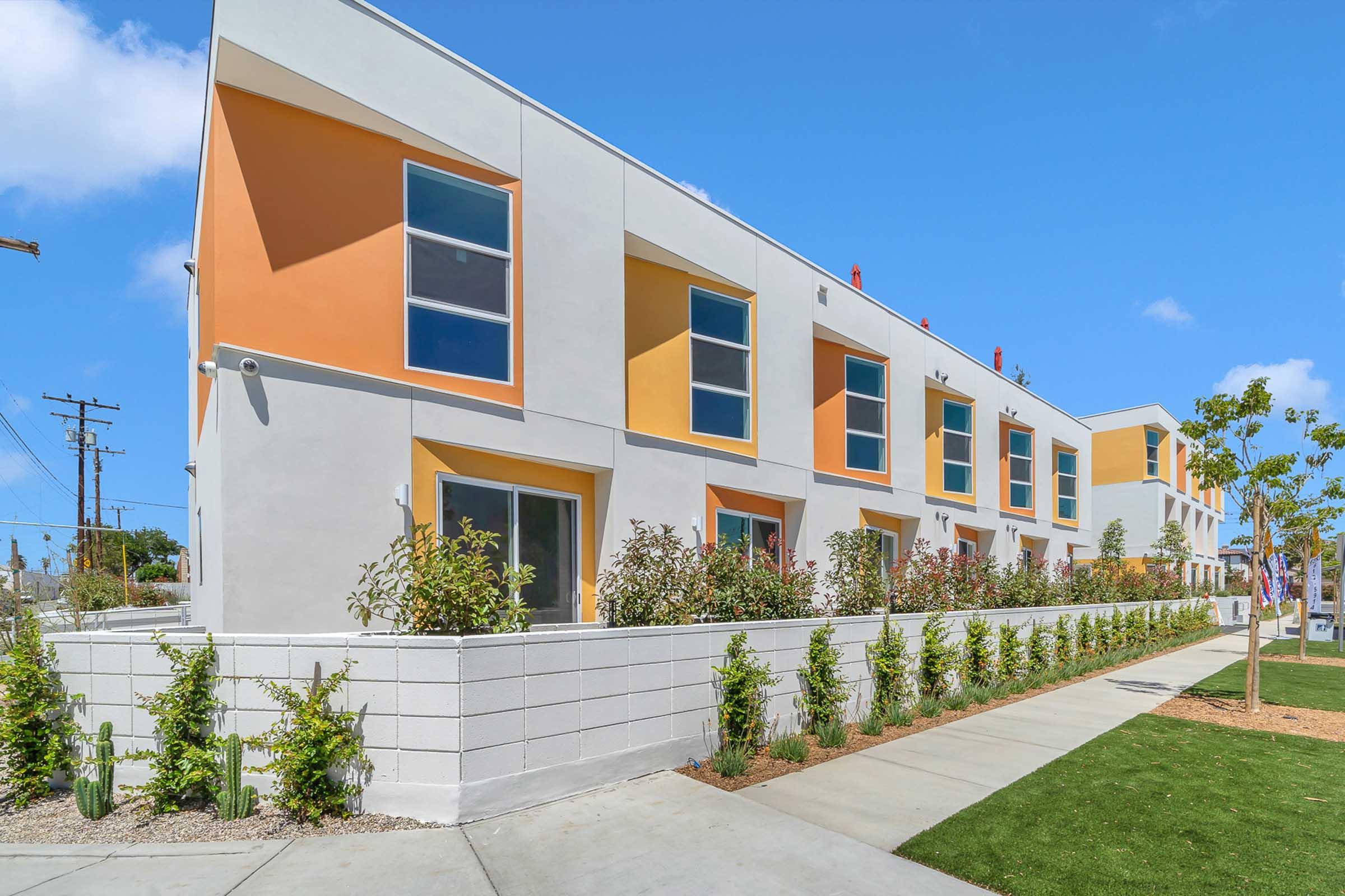 A modern residential building with a white exterior and colorful orange accents. The structure features several large windows and is surrounded by landscaped green areas, including small shrubs and grass. A pathway leads along the front of the building, enhancing its inviting appearance.