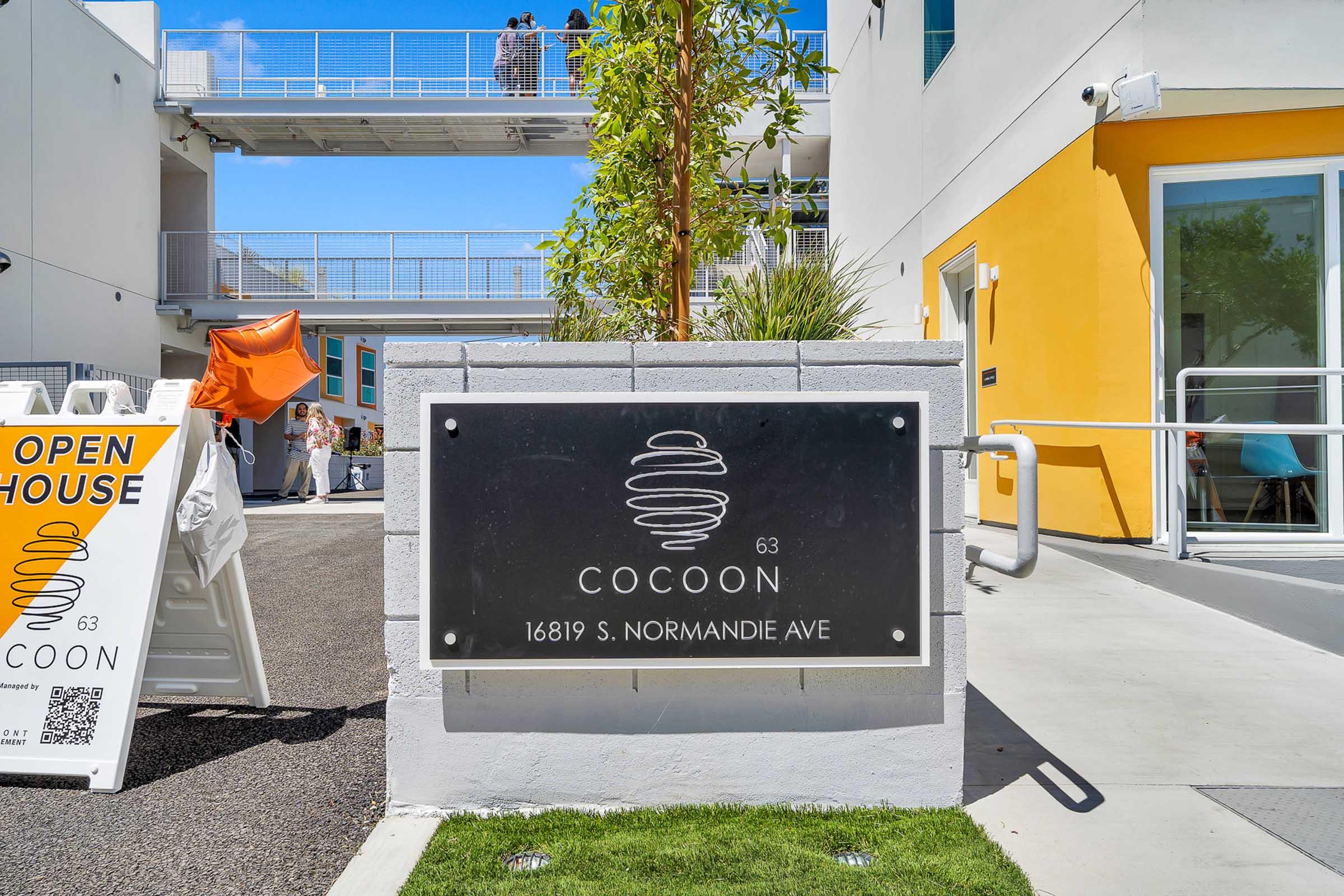 Sign for Cocoon at 16819 S. Normandie Ave, featuring a modern design with a swirl logo. An open house banner is positioned nearby with decorative orange balloons. The building has a contemporary architectural style, with a view of a second story and bright yellow accents.