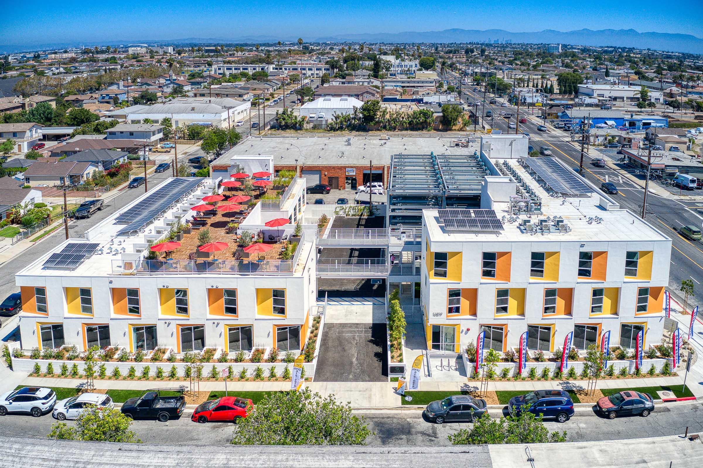 Aerial view of a modern building complex with colorful exterior, featuring a rooftop patio with red umbrellas. Surrounding area includes additional buildings and neighborhoods, with a clear blue sky in the background. Solar panels are visible on the rooftops, indicating a focus on sustainability.