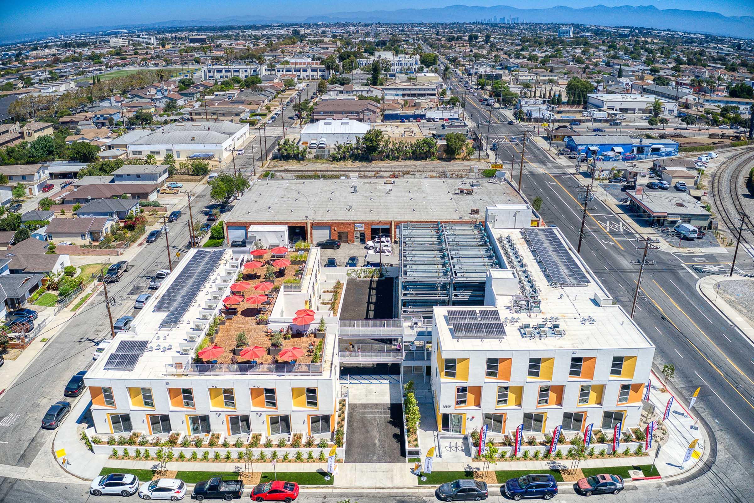 Aerial view of a modern building complex with solar panels on the roof, surrounded by residential areas and streets. The complex features landscaped terraces and outdoor seating with red umbrellas. In the background, there are multiple homes and distant mountains under a clear blue sky.