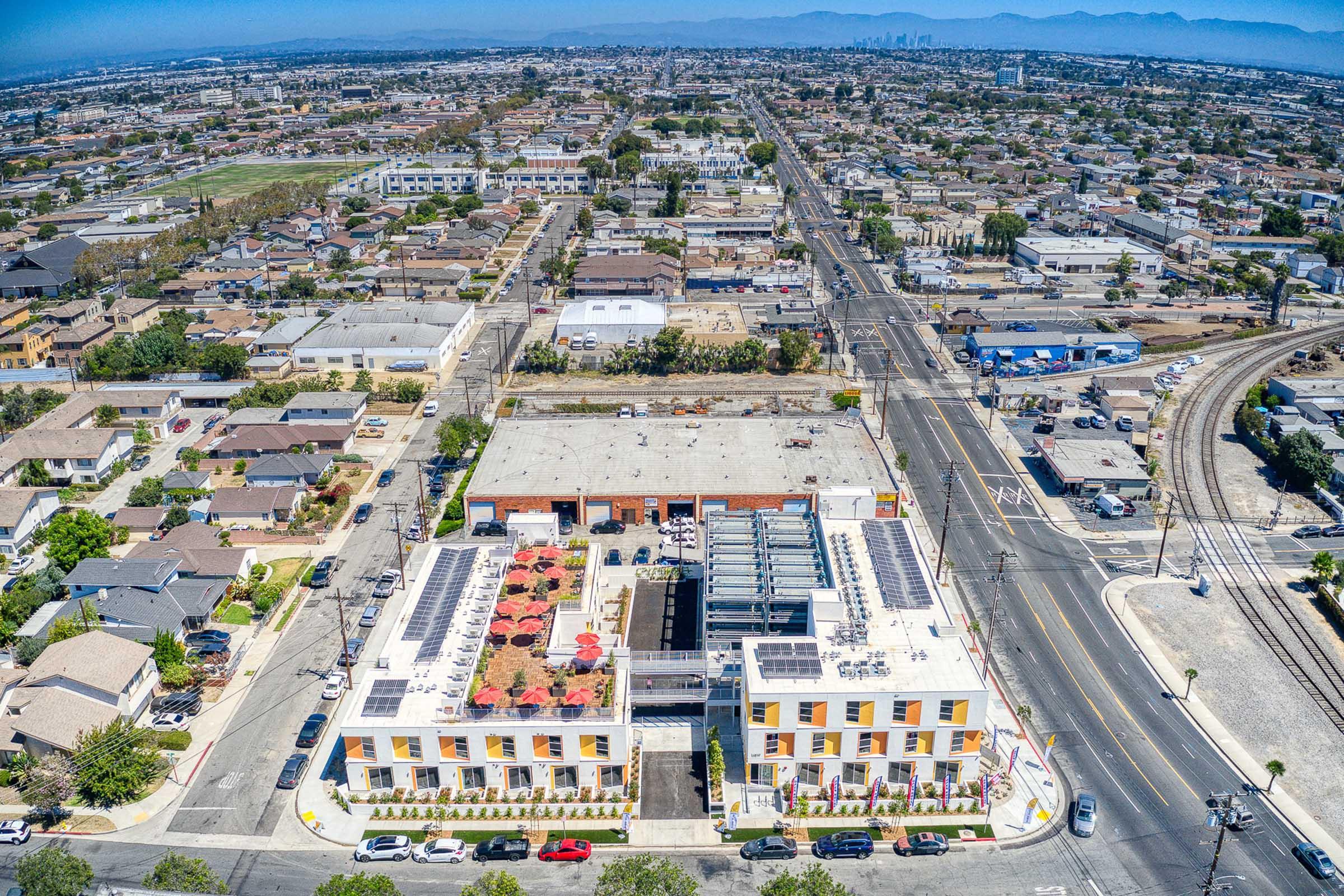 Aerial view of a suburban area featuring residential homes, streets, and a multi-story building with a rooftop terrace and solar panels. Surrounding the building are parking spaces and outdoor seating areas. In the distance, a city skyline is visible against a clear blue sky.