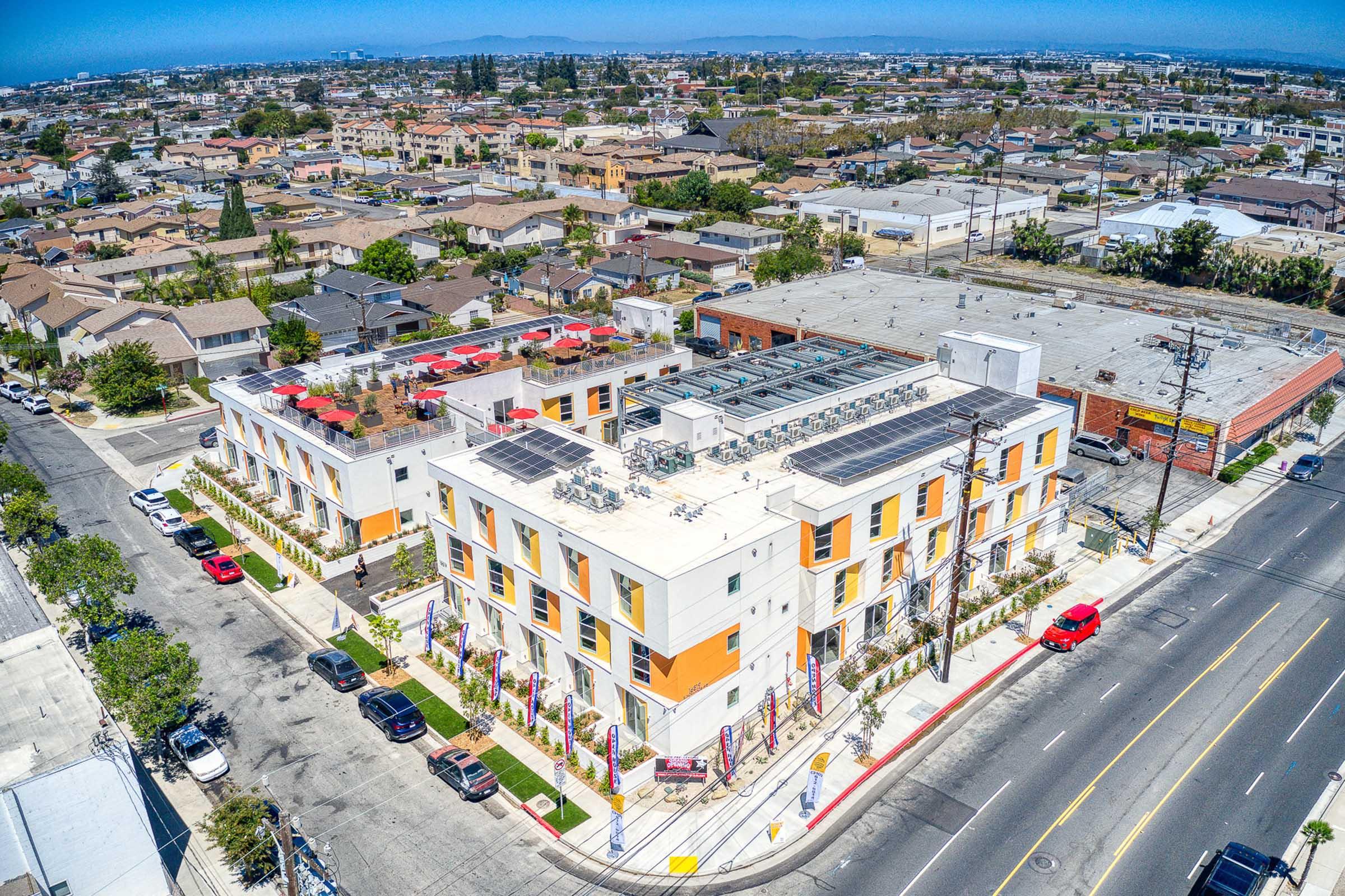 Aerial view of a modern residential building featuring solar panels on the roof, surrounded by green landscaping. The neighborhood consists of sparse urban housing with roads and parked cars visible. The scene is bright and sunny, showcasing a mix of commercial and residential areas in the background.