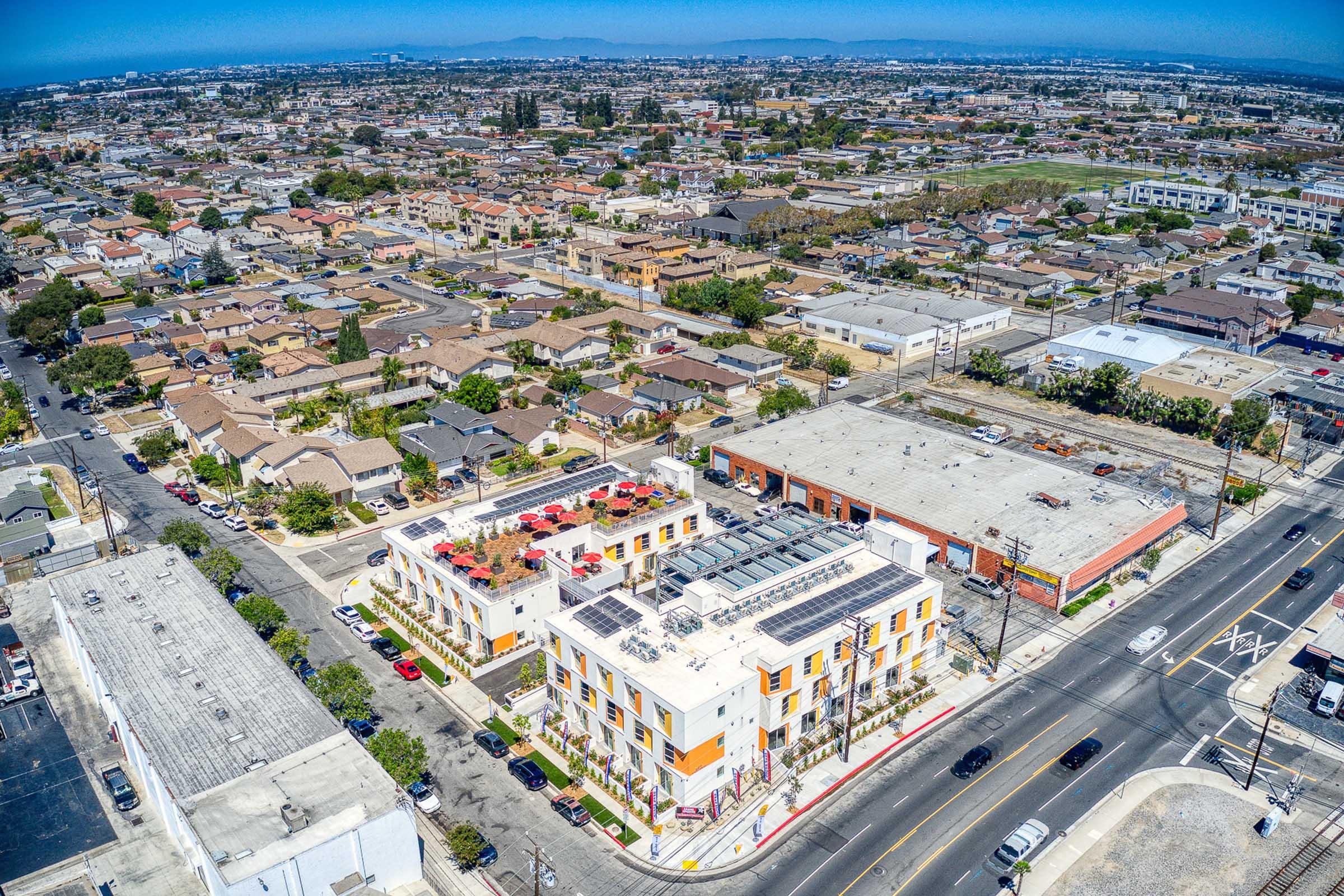 Aerial view of a suburban area featuring a mix of residential homes and commercial buildings. In the foreground, a modern multi-story building with solar panels on the roof stands out, surrounded by streets and parking lots. The background shows a densely populated neighborhood with various structures and greenery.