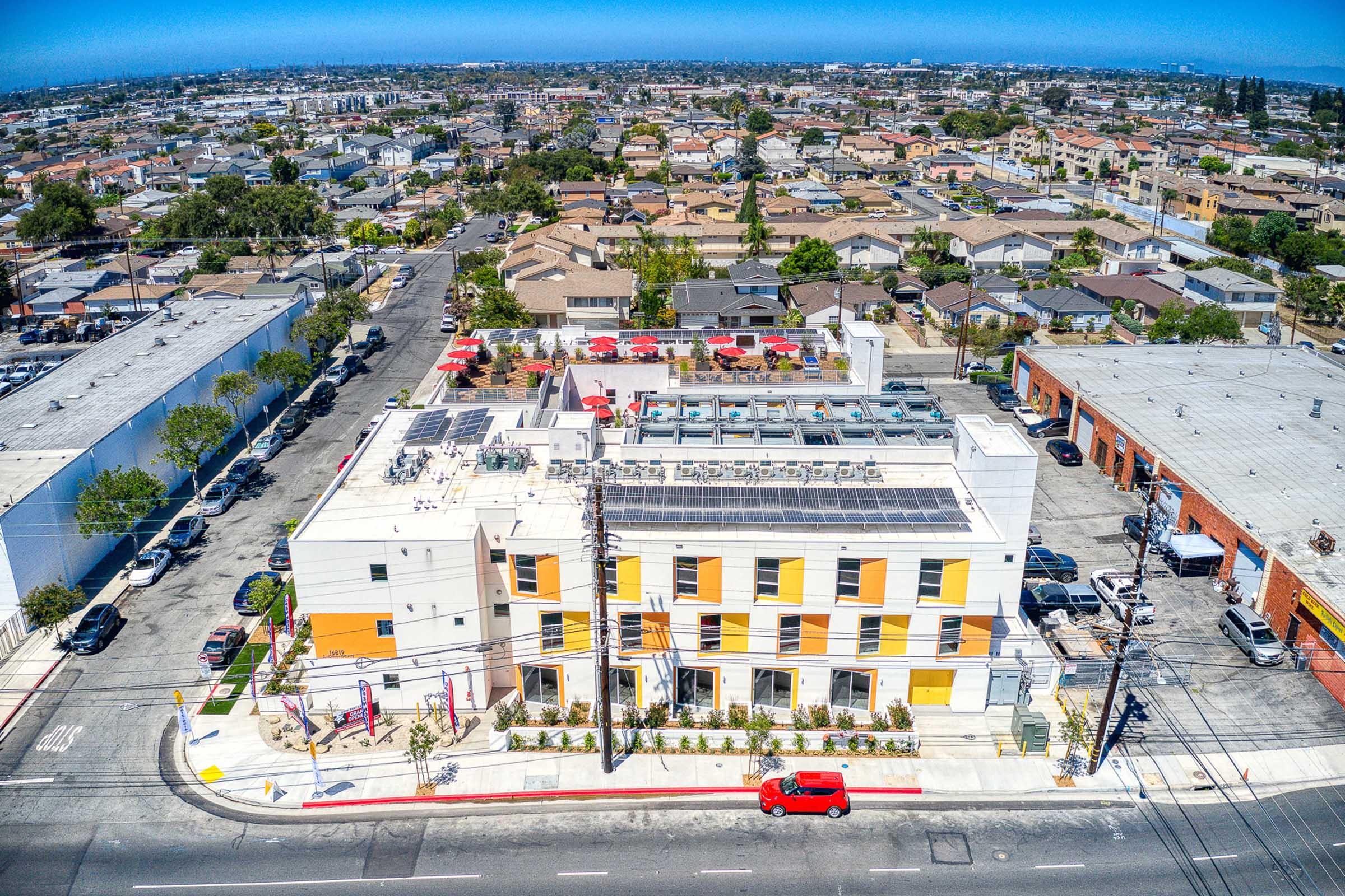 Aerial view of a modern multicolored building with solar panels on the roof, surrounded by residential areas and commercial buildings. The street in front features parked cars, and the surrounding area showcases a mix of rooftops and greenery against a clear blue sky.