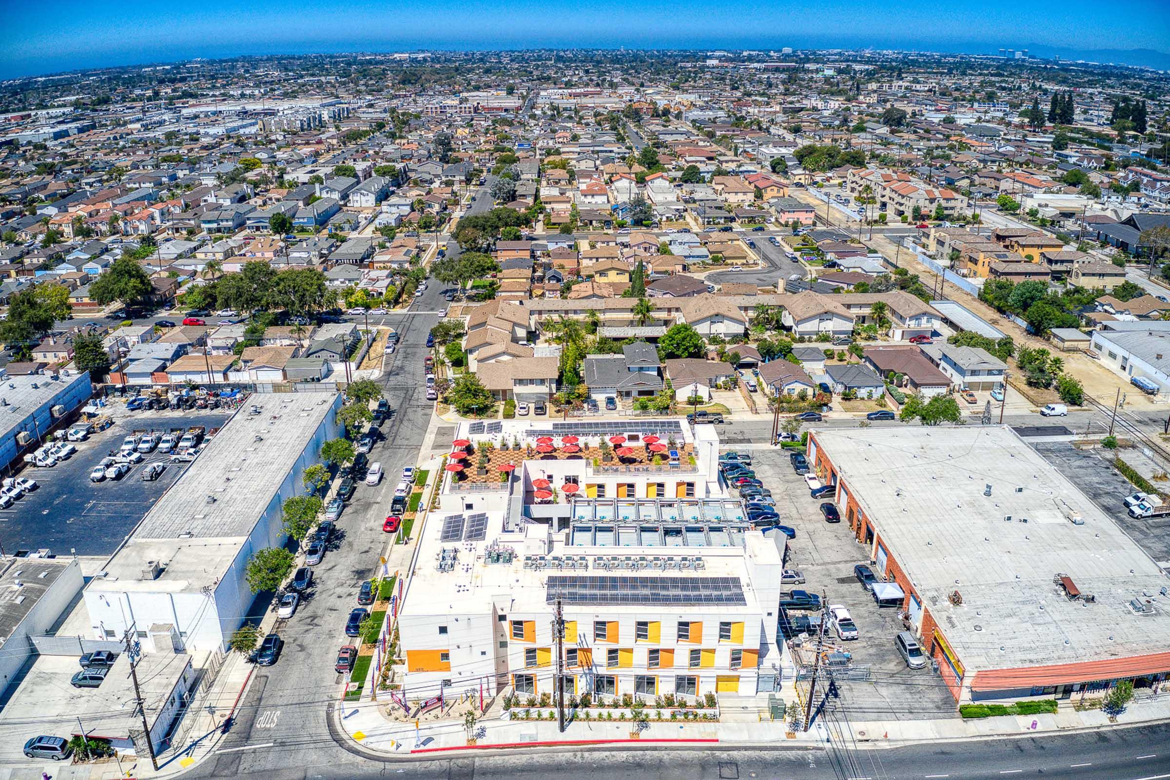Aerial view of a suburban area featuring a mix of residential houses and commercial buildings. In the foreground, a colorful multi-story building with a rooftop terrace is visible, surrounded by parking lots and roads. The background shows a sprawling neighborhood with a clear blue sky overhead.