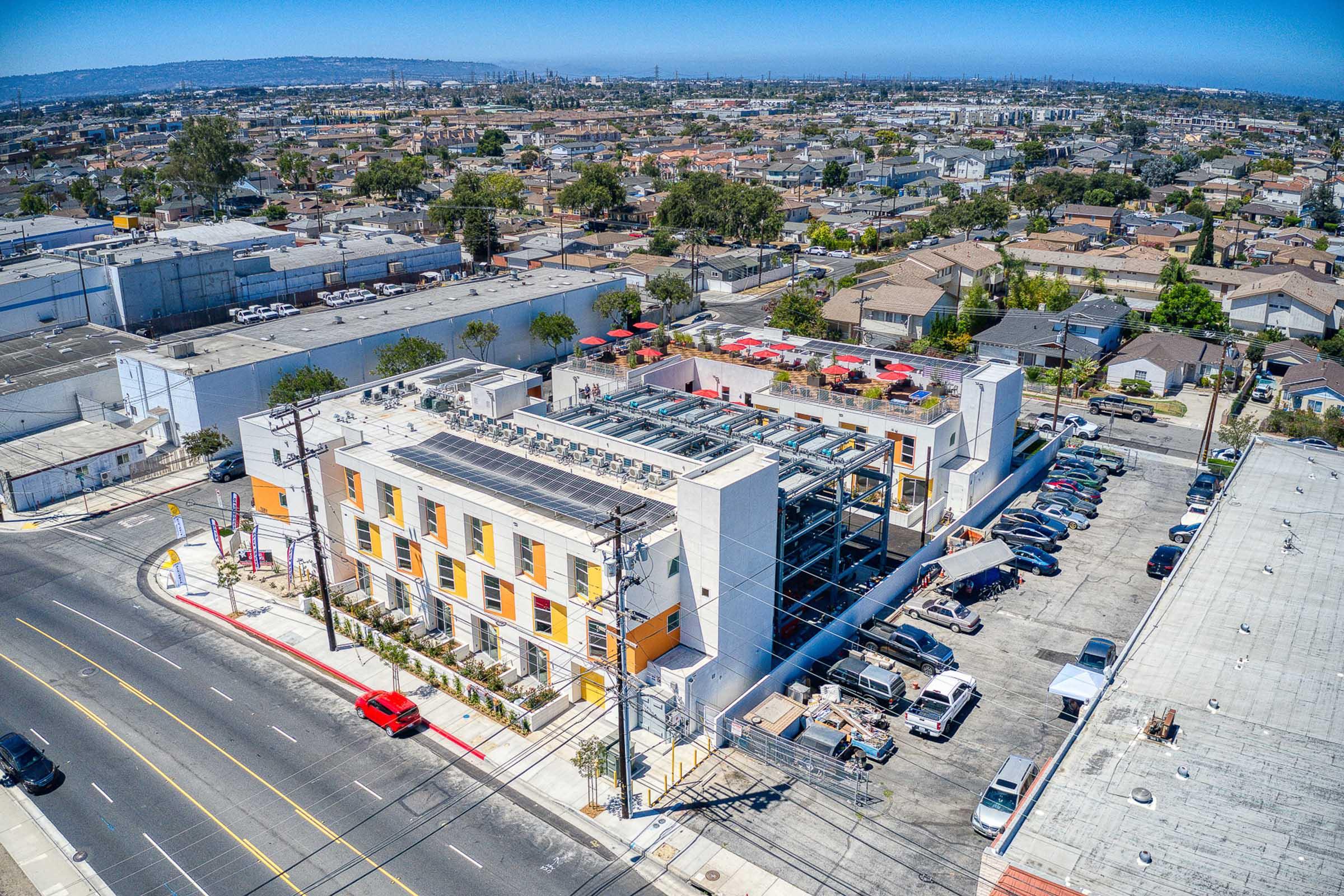 Aerial view of a modern multi-story building with a rooftop solar panel installation, surrounded by a parking lot filled with vehicles. Nearby, a mix of residential and commercial buildings stretches into the distance under a clear blue sky.