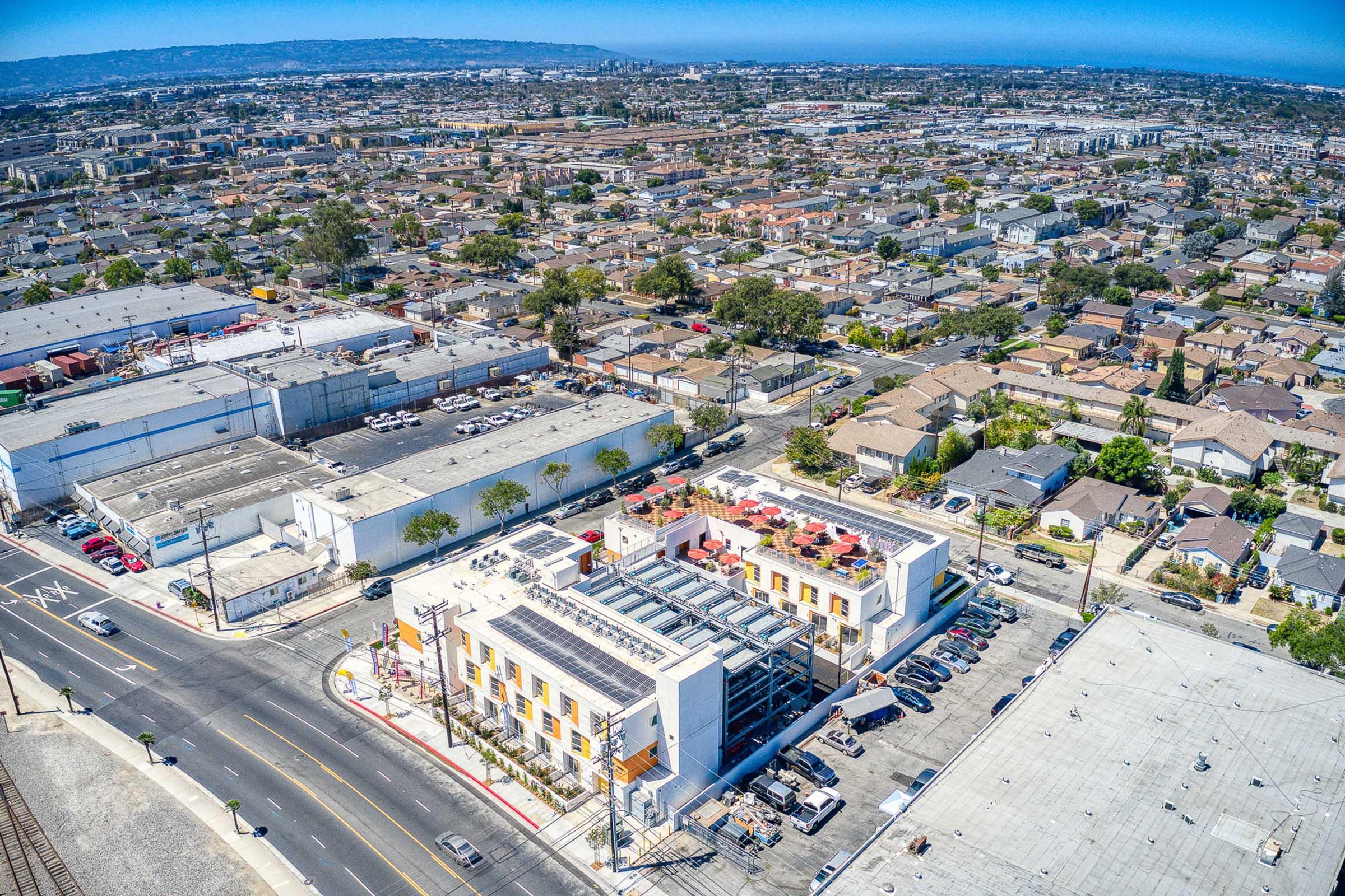 Aerial view of an urban area featuring a modern commercial building with a rooftop patio, surrounded by a variety of residential homes and warehouses. The landscape includes roads, parking lots, and a clear blue sky, illustrating a bustling neighborhood setting.