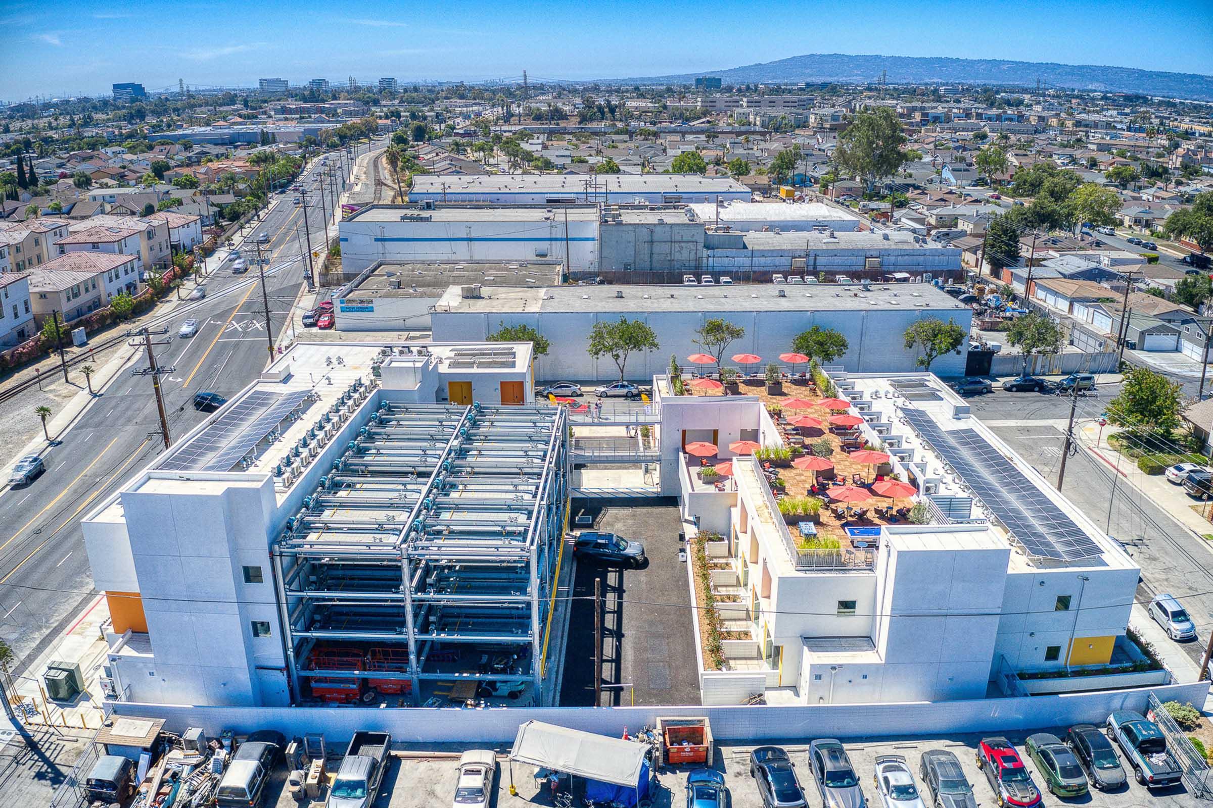 Aerial view of a modern building with solar panels on the roof, surrounded by a parking lot and outdoor seating areas with red umbrellas. The building is part of a larger urban landscape with residential areas and streets visible in the background, showcasing a blend of architecture and city life.