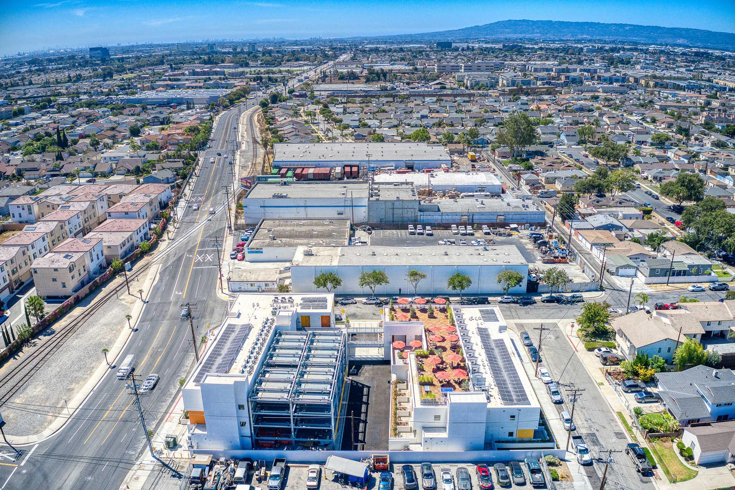 Aerial view of a suburban area featuring rows of residential buildings, a commercial building with a rooftop garden, and a street lined with cars. In the background, industrial structures and a distant skyline can be seen under a clear blue sky.