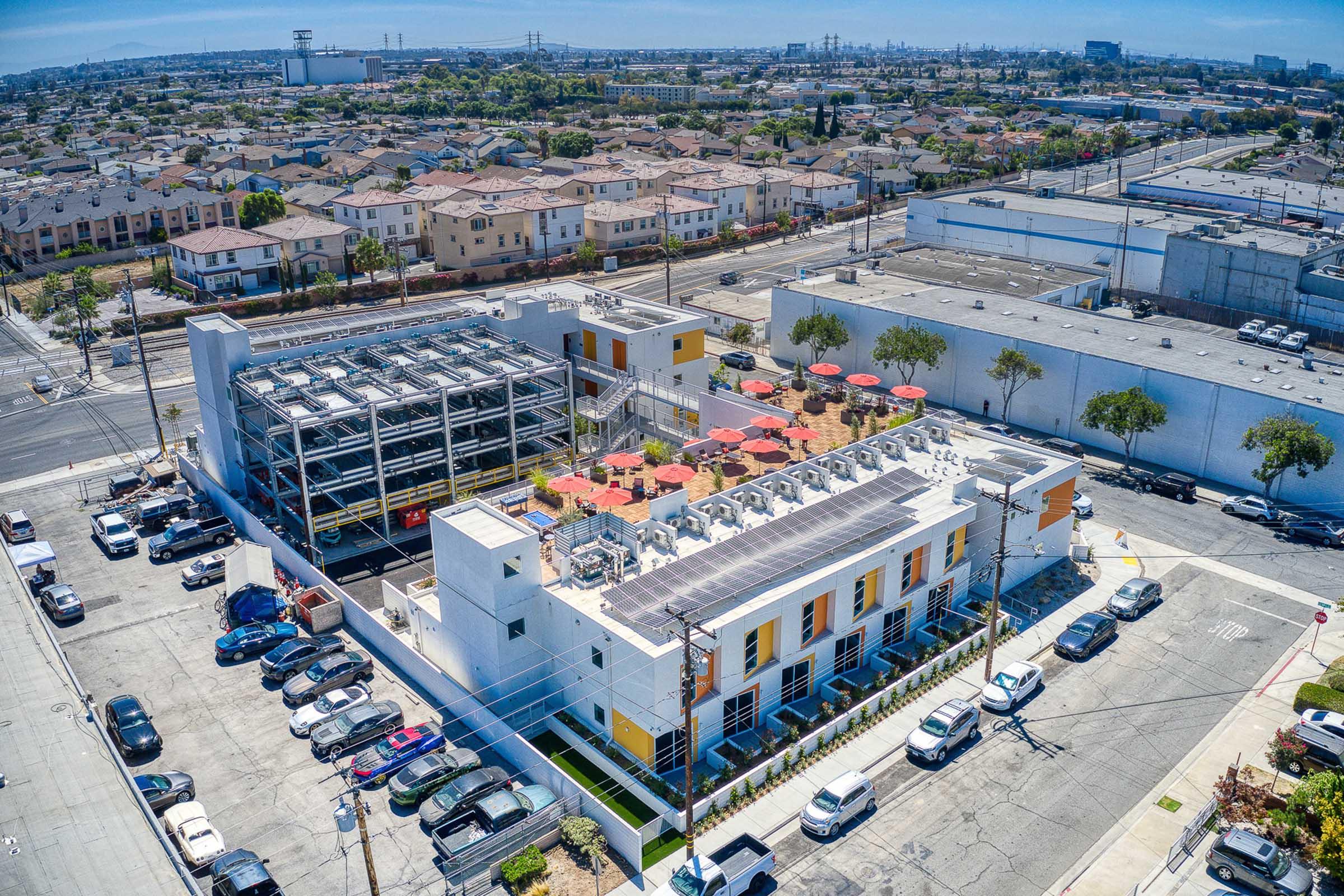 Aerial view of a modern building with solar panels on the roof, surrounded by parked cars and residential areas. The structure features outdoor seating areas and is situated in an urban setting with industrial buildings in the background. Bright blue sky overhead.