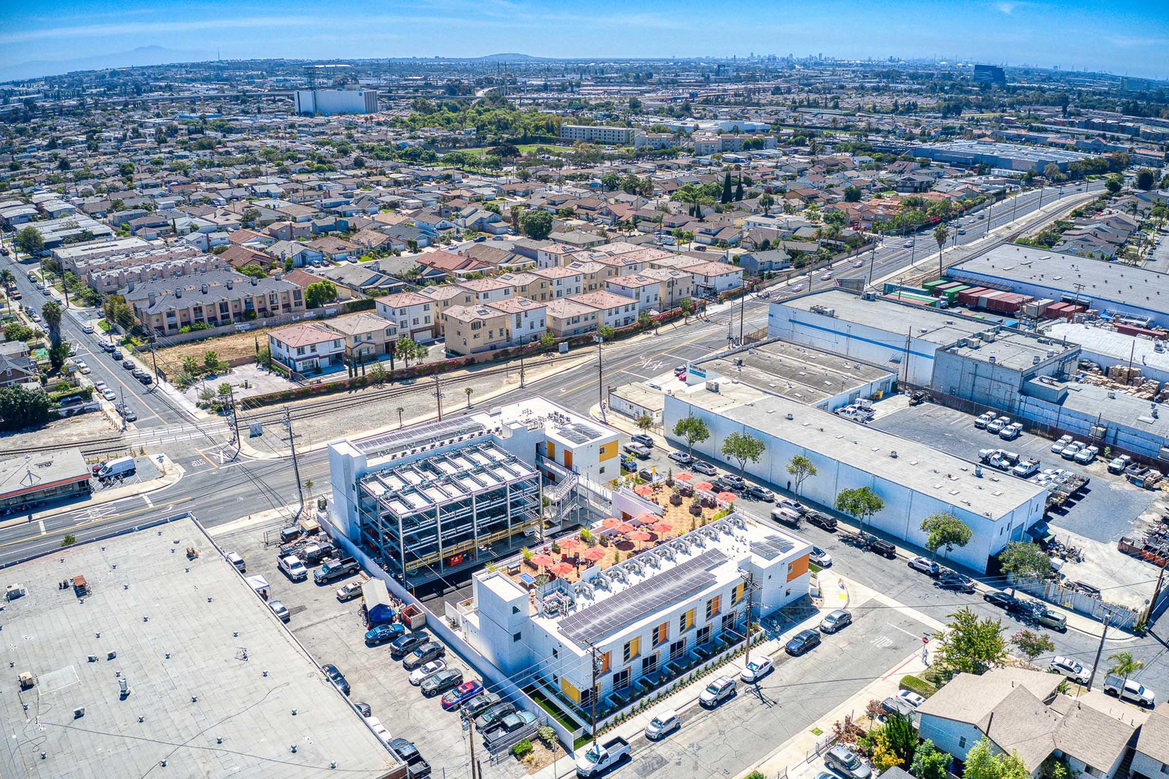 Aerial view of a suburban area featuring a mix of residential buildings and commercial structures. In the foreground, a multi-story building with a rooftop terrace and parking lot is visible, surrounded by several single-family homes and warehouses in the background. The landscape is dotted with greenery and roads.
