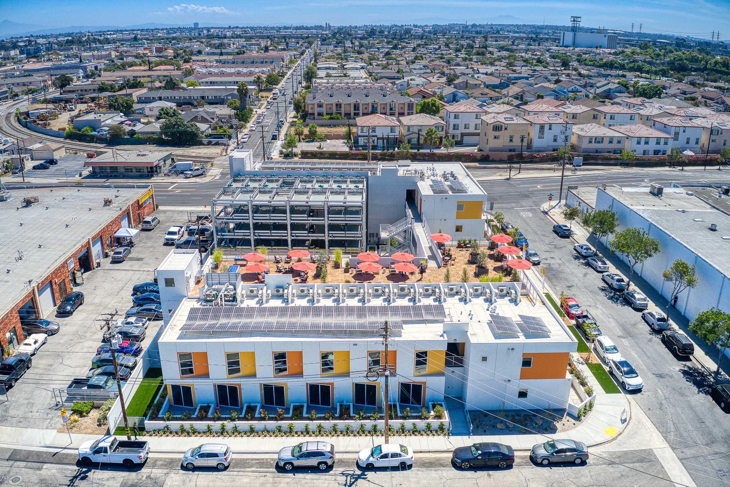 Aerial view of a modern building with solar panels and a rooftop garden, surrounded by residential and commercial areas. The building features a landscaped area with umbrellas, and various parking spaces, set against a backdrop of city streets and homes. Industry and infrastructure visible in the distance.