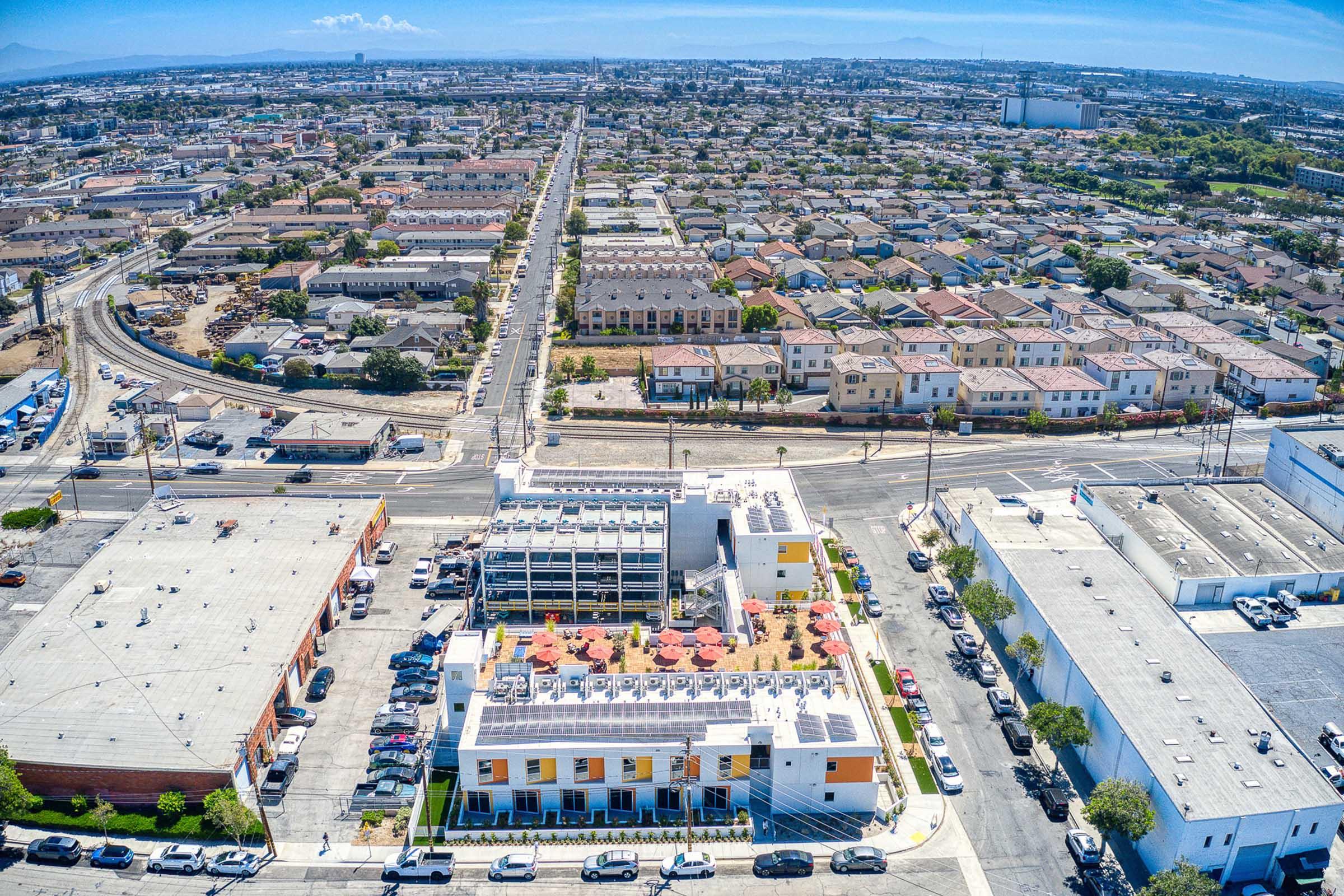 Aerial view of a residential area featuring a mix of low-rise buildings, houses, and a parking lot. In the foreground, a modern multi-story building with rooftop seating and colorful umbrellas is visible. The background shows a suburban neighborhood with numerous houses and streets stretching into the distance.
