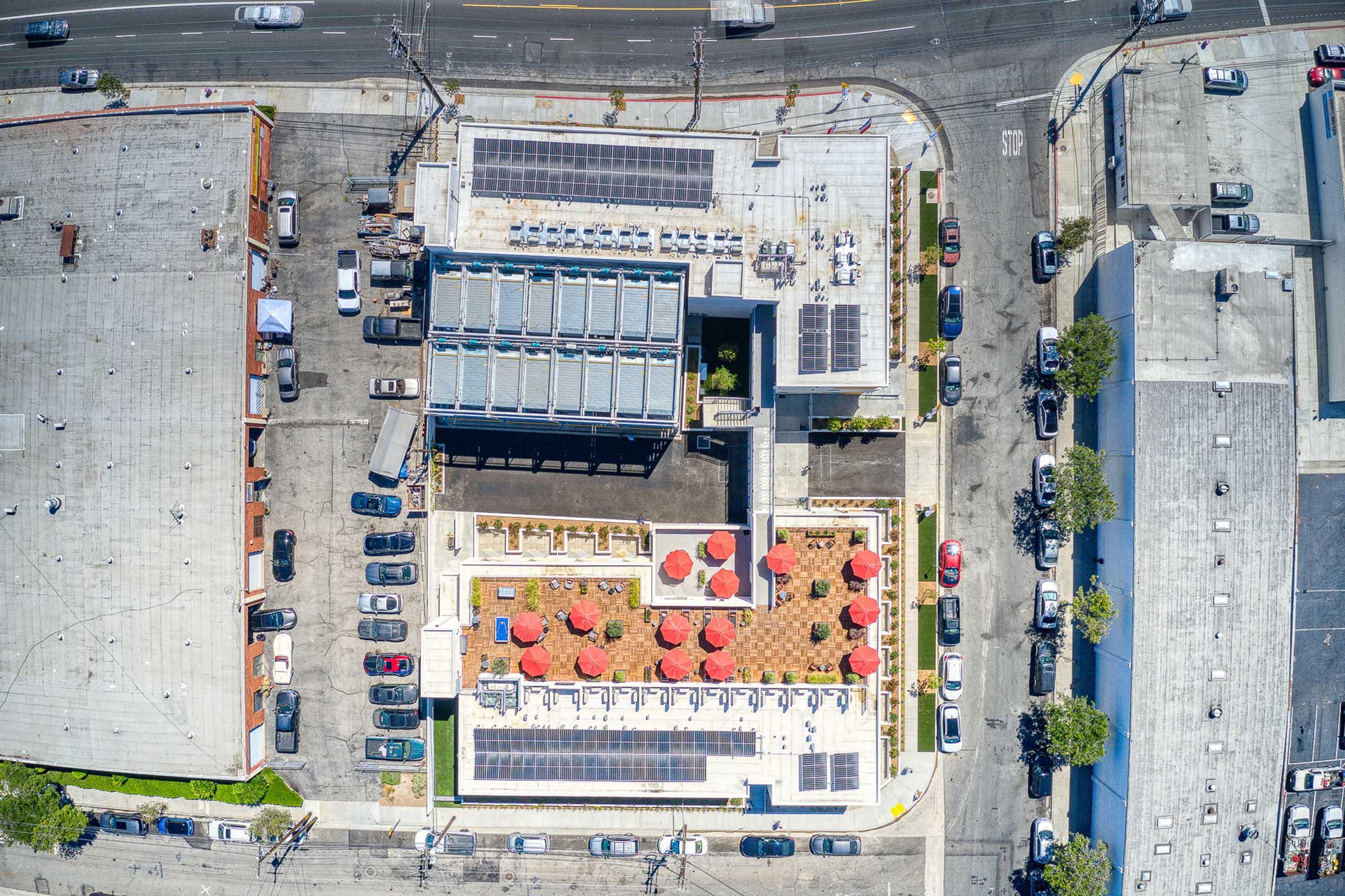 Aerial view of a modern building with a rooftop terrace featuring solar panels, surrounded by a parking area. The terrace has red umbrellas over tables, while nearby buildings and a road are visible. The scene captures urban development with green energy solutions.