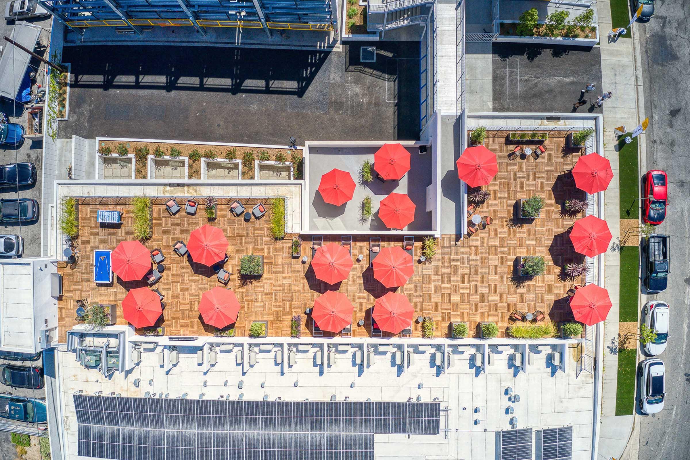 Aerial view of a rooftop terrace featuring several red umbrellas, lounge chairs, and potted plants. The floor is made of wooden tiles, and solar panels are visible on one side. In the background, adjacent buildings and parked cars can be seen.