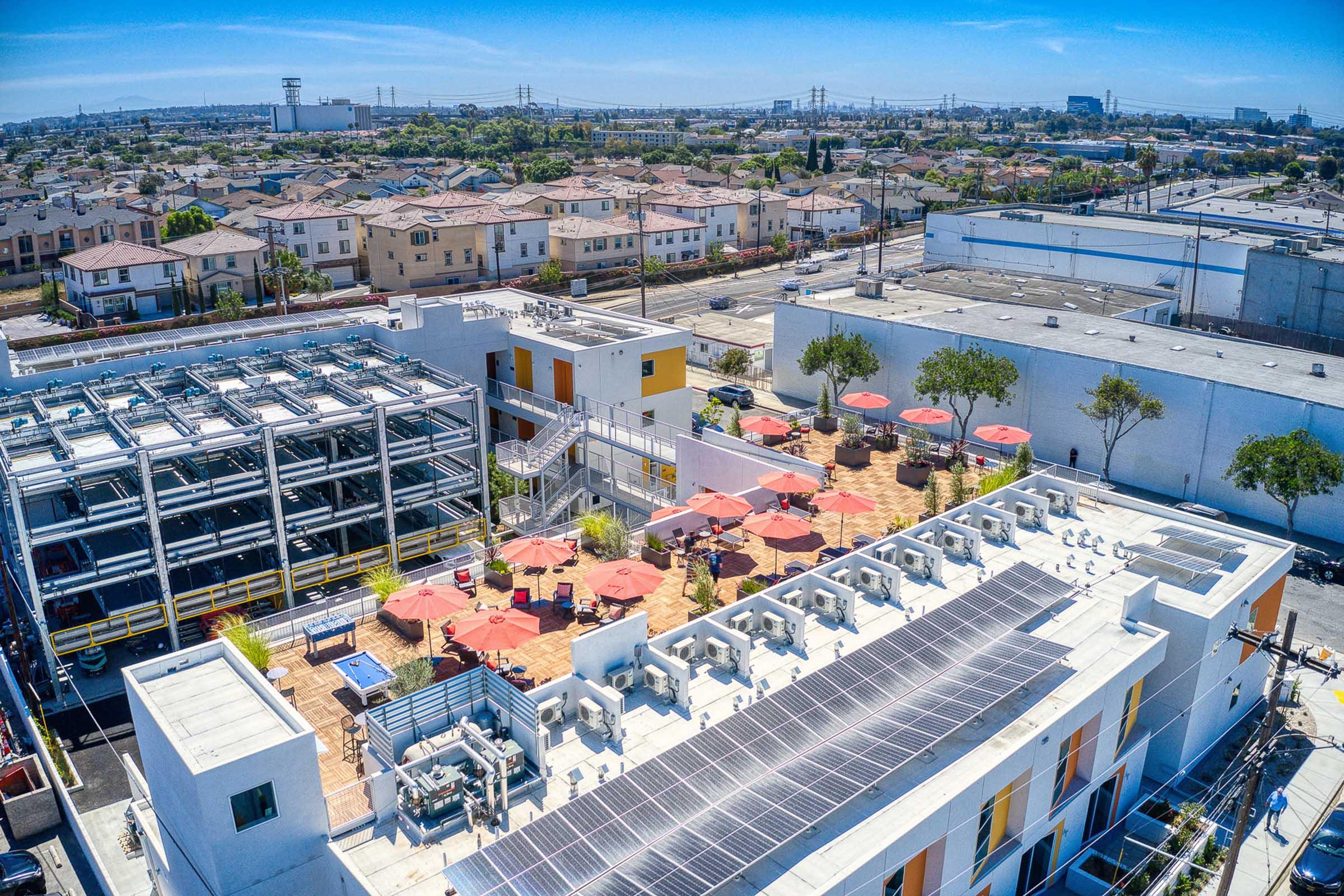 Aerial view of a rooftop space featuring solar panels, landscaped seating areas with red umbrellas, and adjacent buildings. In the background, a residential area with houses, power lines, and industrial structures can be seen under a clear blue sky.