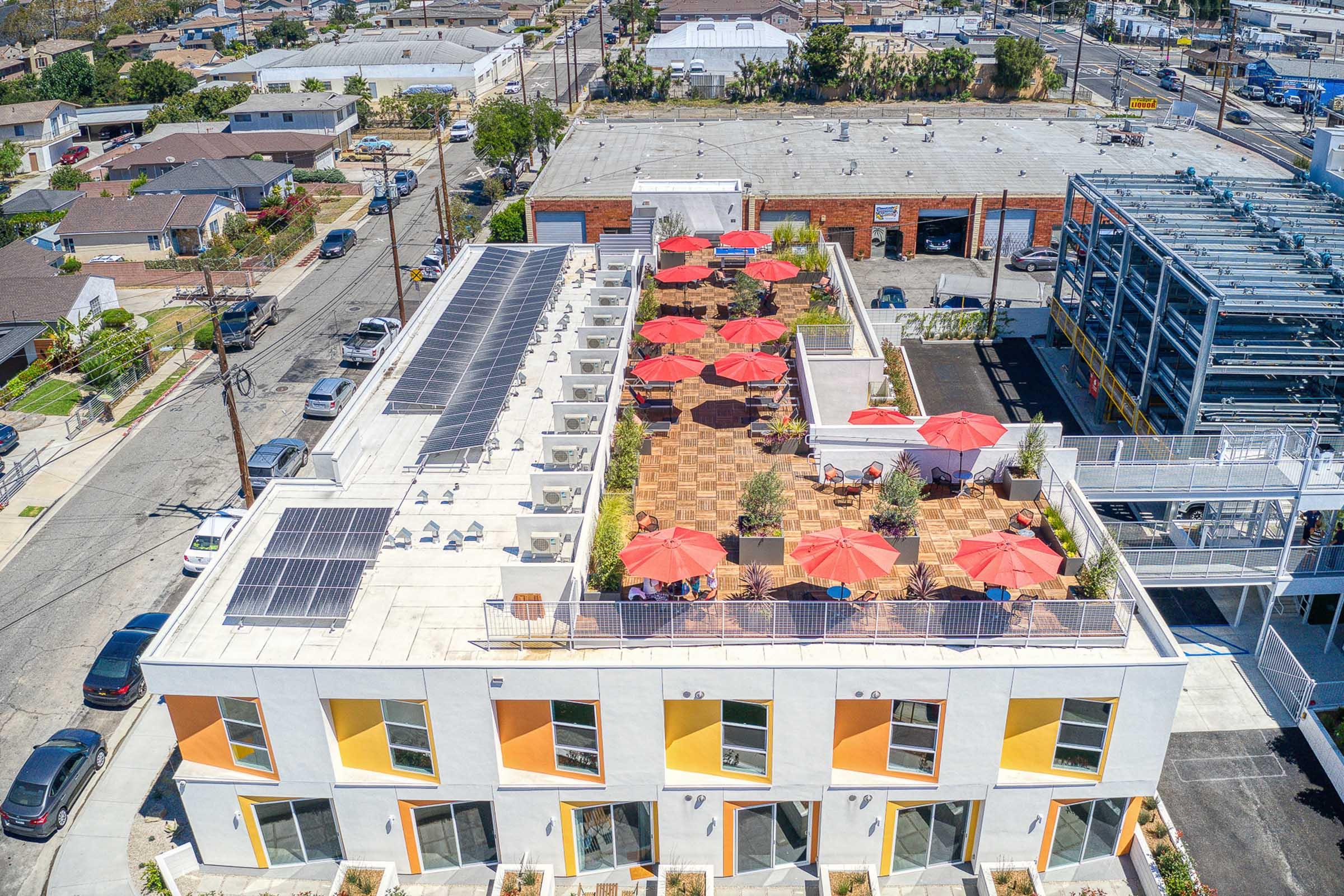 Aerial view of a building rooftop featuring solar panels, outdoor seating areas with red umbrellas, and a landscaped terrace. Surrounding buildings and streets are visible, showcasing a mix of residential and commercial properties. Bright sunlight illuminates the scene.