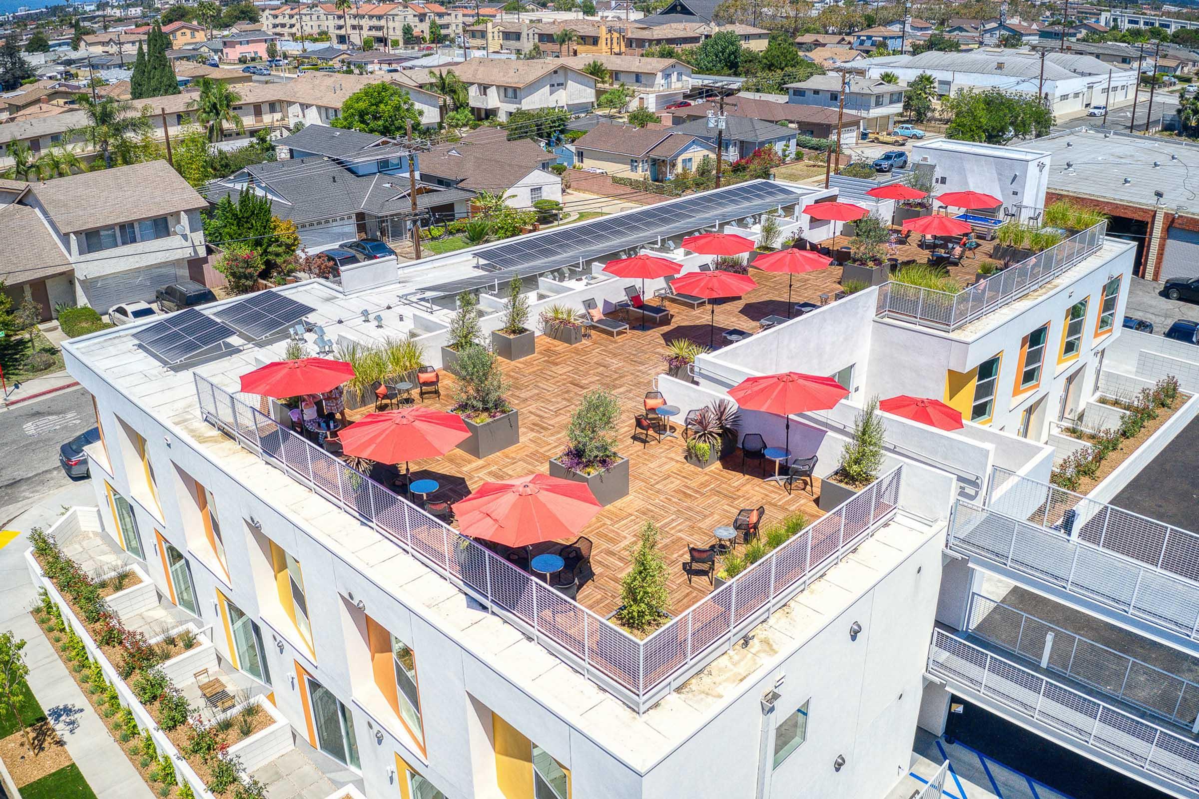 Aerial view of a multi-story building featuring a rooftop terrace adorned with red umbrellas, small tables, and green plants. Solar panels are visible on the roof, and residential neighborhoods extend into the background, showcasing a clear sky and a mix of buildings in the area.