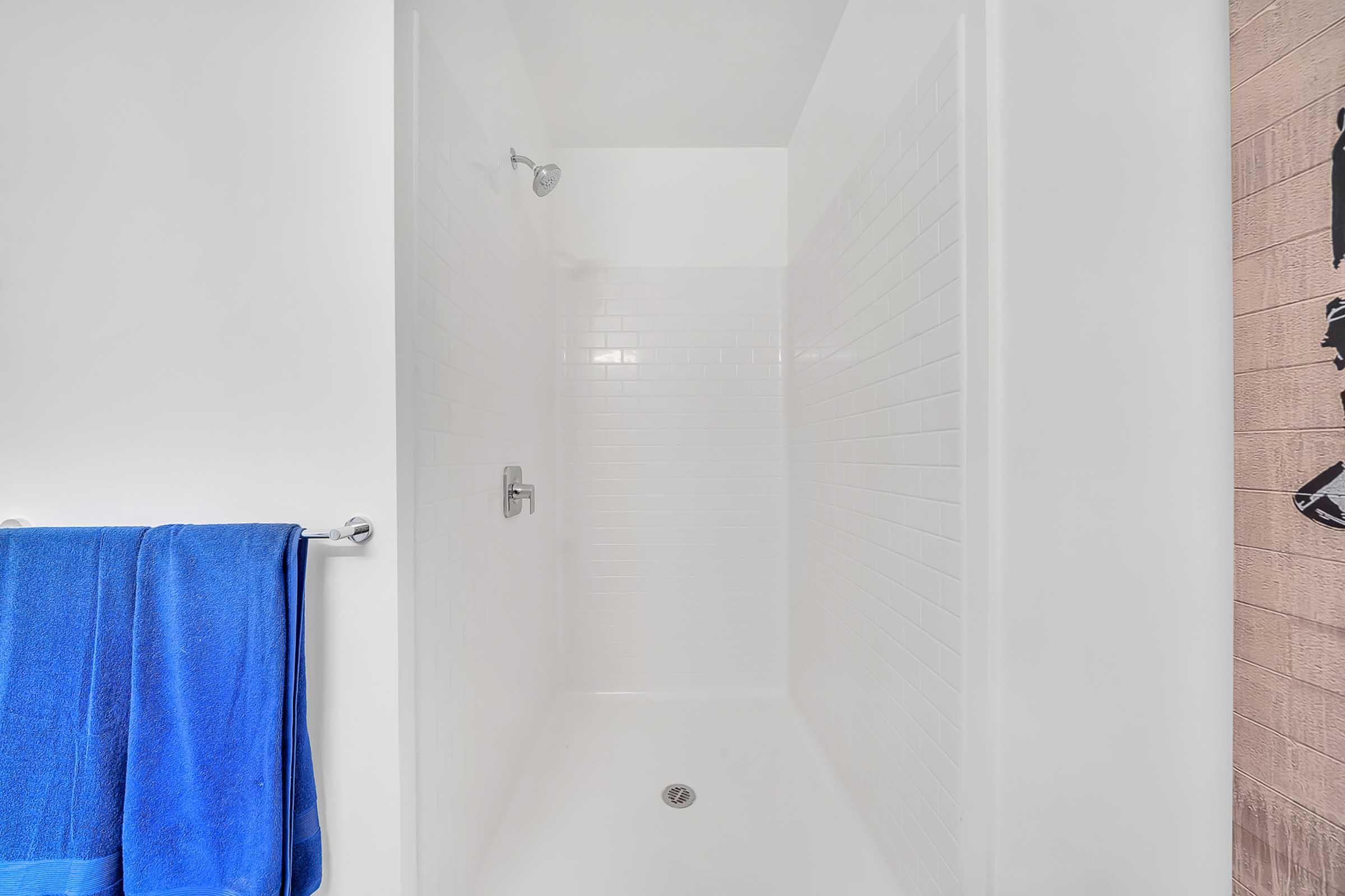 A clean, modern shower stall with white tile walls and a frosted glass shower door. A blue towel hangs on a chrome towel bar. The floor is smooth and featureless, creating a minimalist aesthetic.