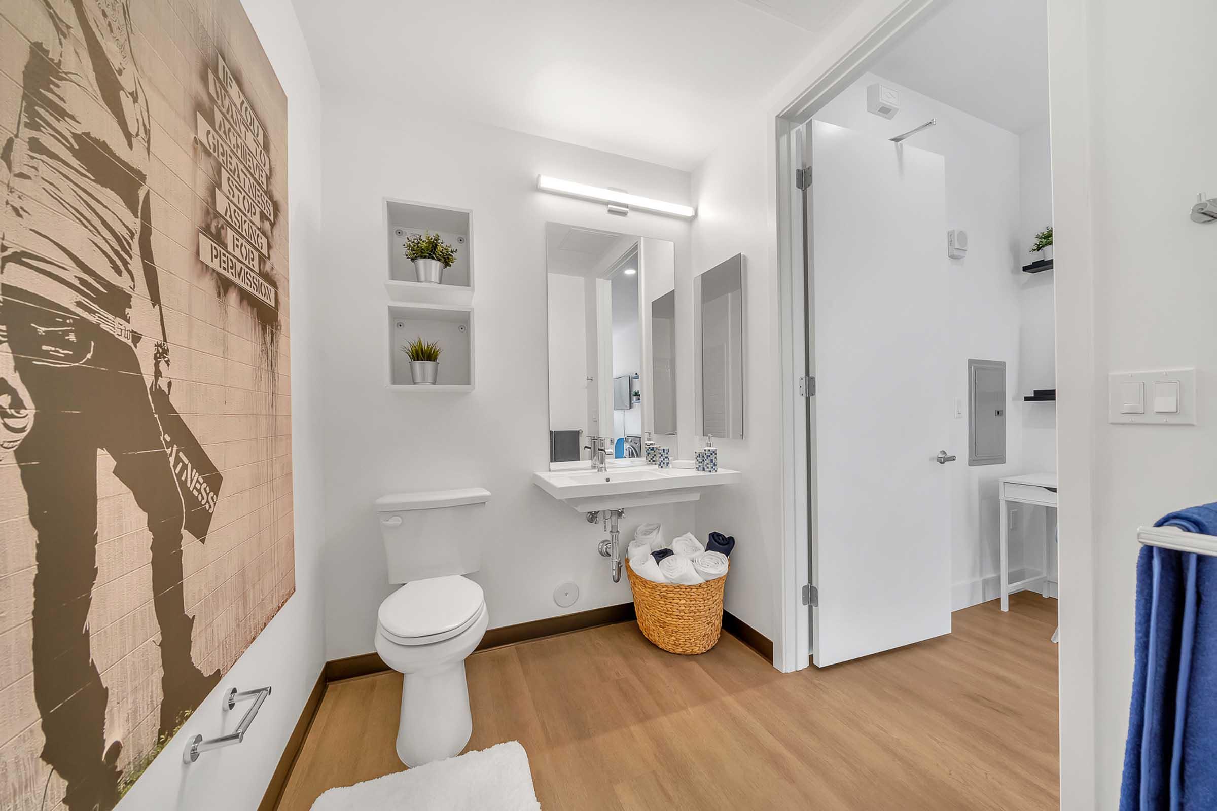 Modern bathroom featuring a stylish sink with a mirror, a toilet, and decorative shelving. A basket for laundry is on the floor, and a wall art piece is visible. The overall design is clean and contemporary with light colors and wood flooring. A towel hangs on the door.