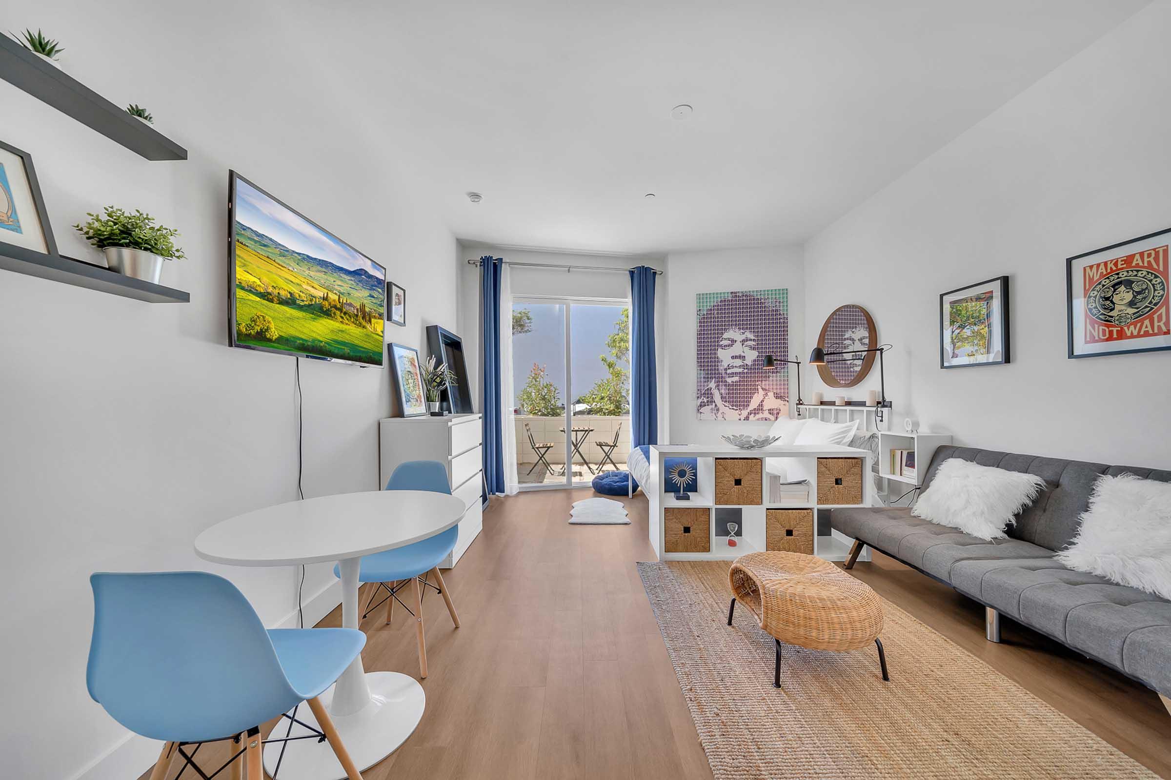 A cozy living space featuring a grey sofa, a round dining table with blue chairs, and a white TV stand. Decor includes wall art, plants on shelves, and a light-colored area rug. Large windows with blue curtains offer a view of the outdoors, creating a bright and inviting atmosphere.