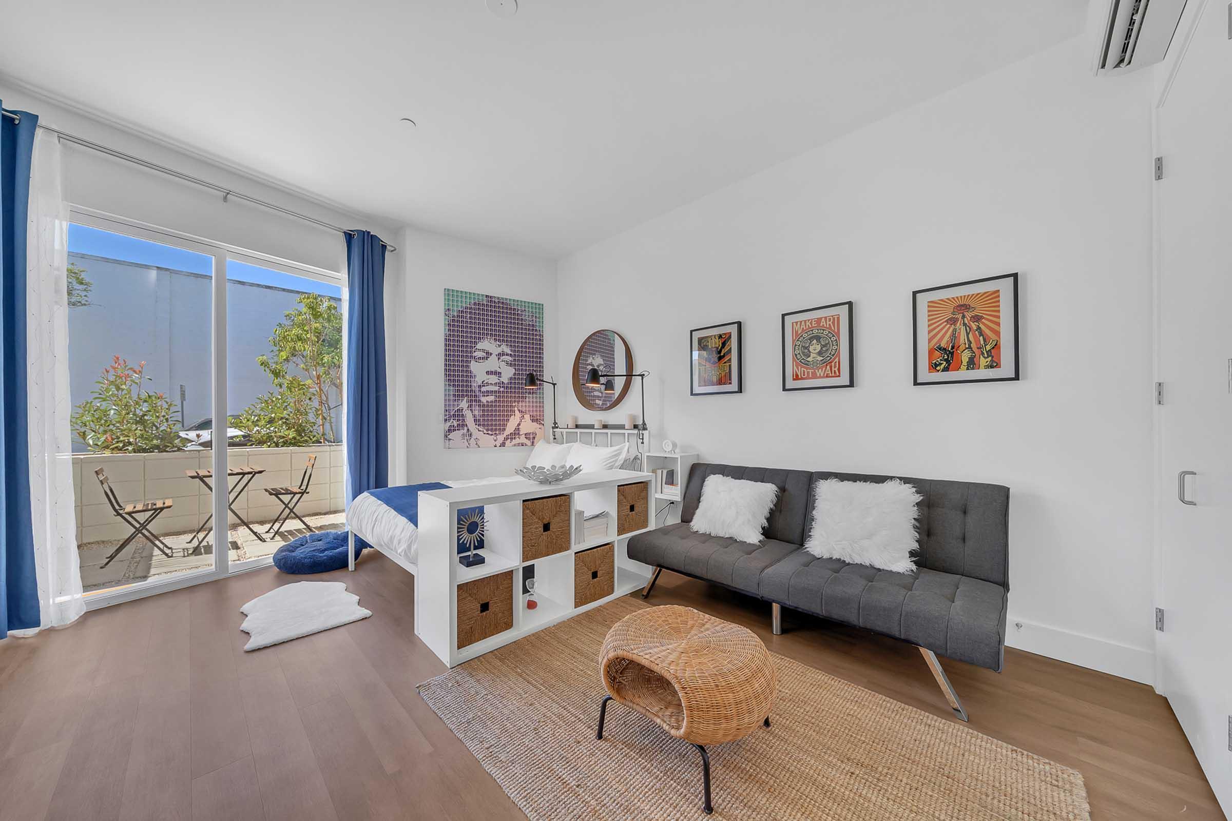 A bright and modern bedroom featuring a large window with blue curtains, a gray sofa with decorative pillows, a wooden storage unit, and art on the walls. A round mirror is above the unit, and a cozy rug is on the floor. A small balcony with outdoor seating is visible outside the window.