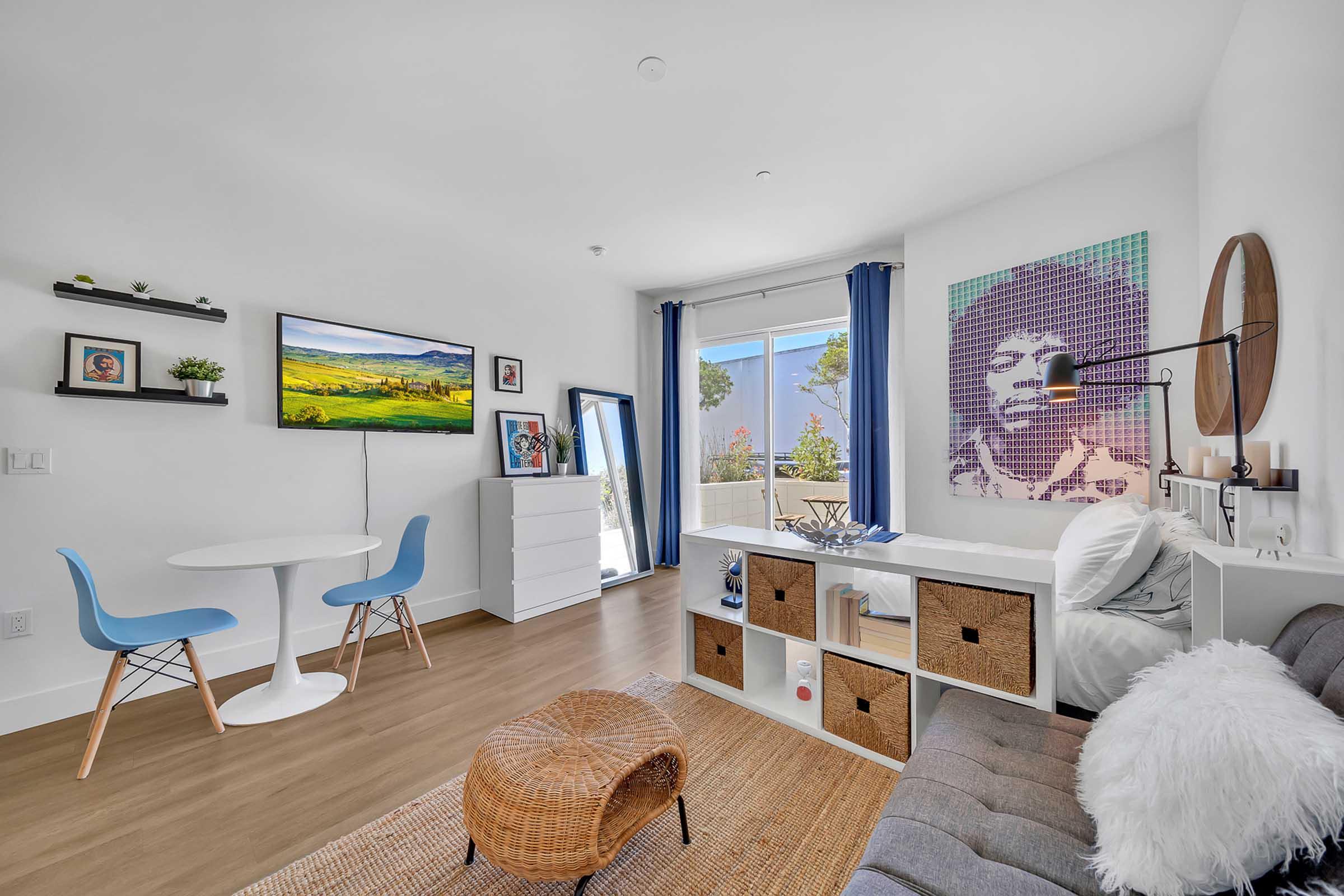 A modern, stylish living space featuring a small dining table with blue chairs, a white dresser, a wall-mounted TV, and a cozy couch. The room is bright with large windows, decorated with a piece of art depicting a musician, and has natural light streaming in, creating a welcoming atmosphere.