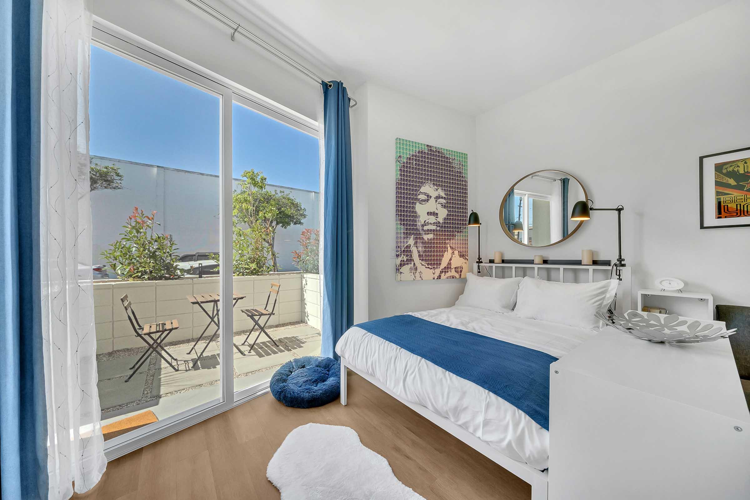 A modern bedroom featuring a large window with sheer curtains and a view of a small balcony with two chairs. The bed is dressed in white linens with a blue throw, and there's a circular mirror above the bed. A colorful piece of art is displayed on the wall, adding a vibrant touch to the room's neutral decor.