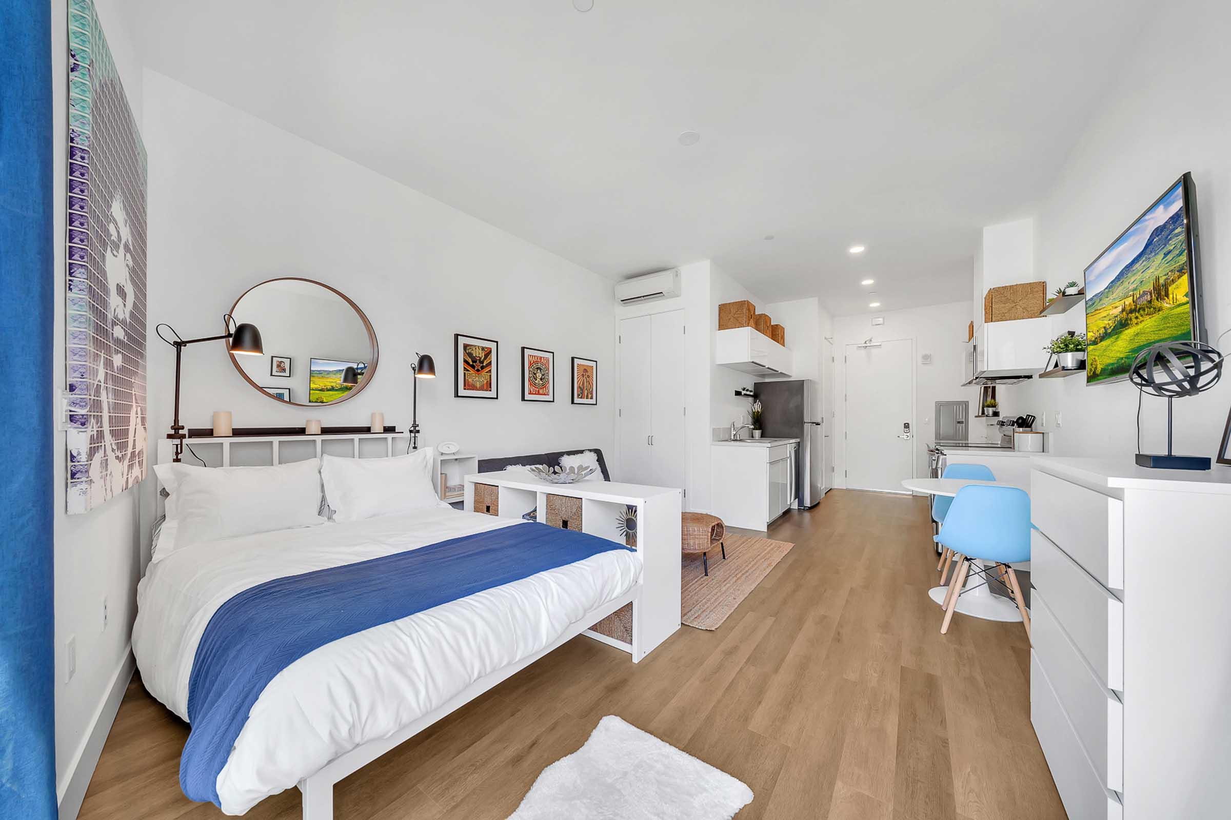 A modern studio apartment featuring a bed with a blue throw, a small dining area, a kitchenette, and decorative artwork on the walls. Natural light brightens the space, which includes wooden flooring and stylish furniture, creating a cozy and inviting atmosphere.