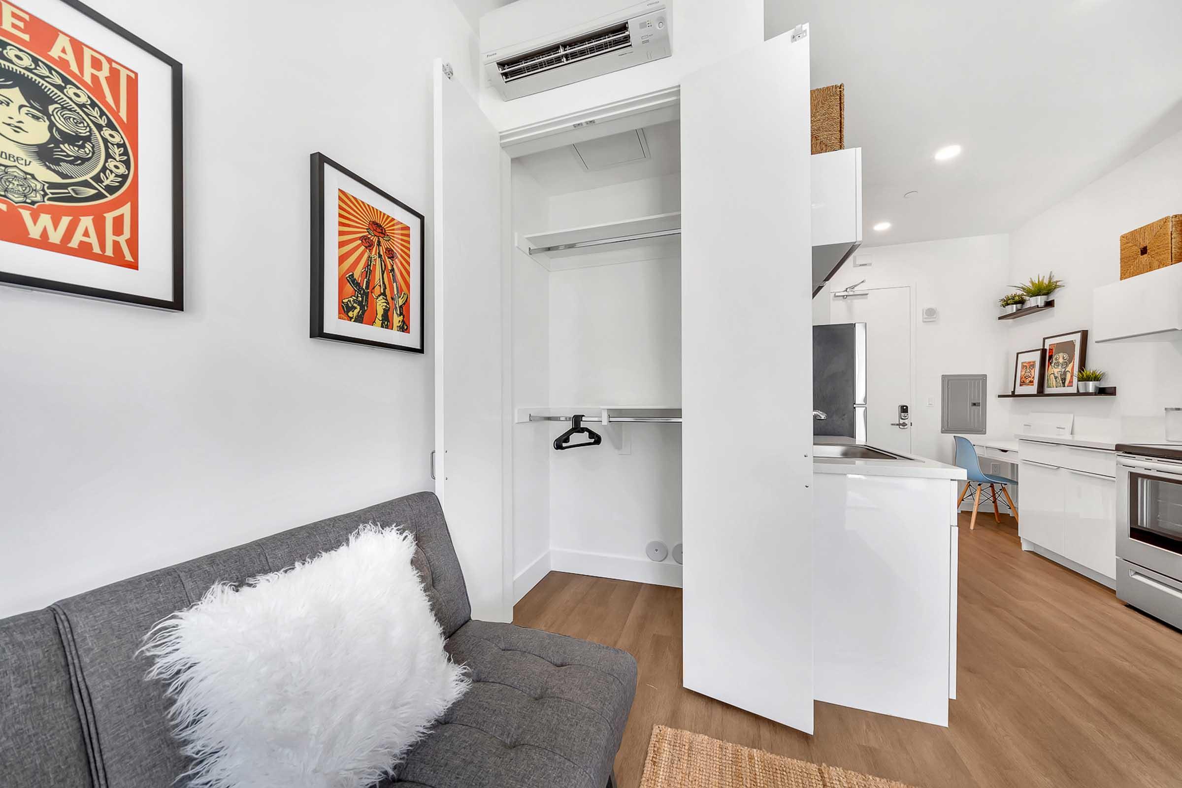 A small, modern living space featuring a gray sofa with a fluffy white throw pillow, a closet with open doors, and framed artwork on the walls. The kitchen area is visible in the background, showcasing a sleek design with light-colored cabinetry and a stainless steel oven.
