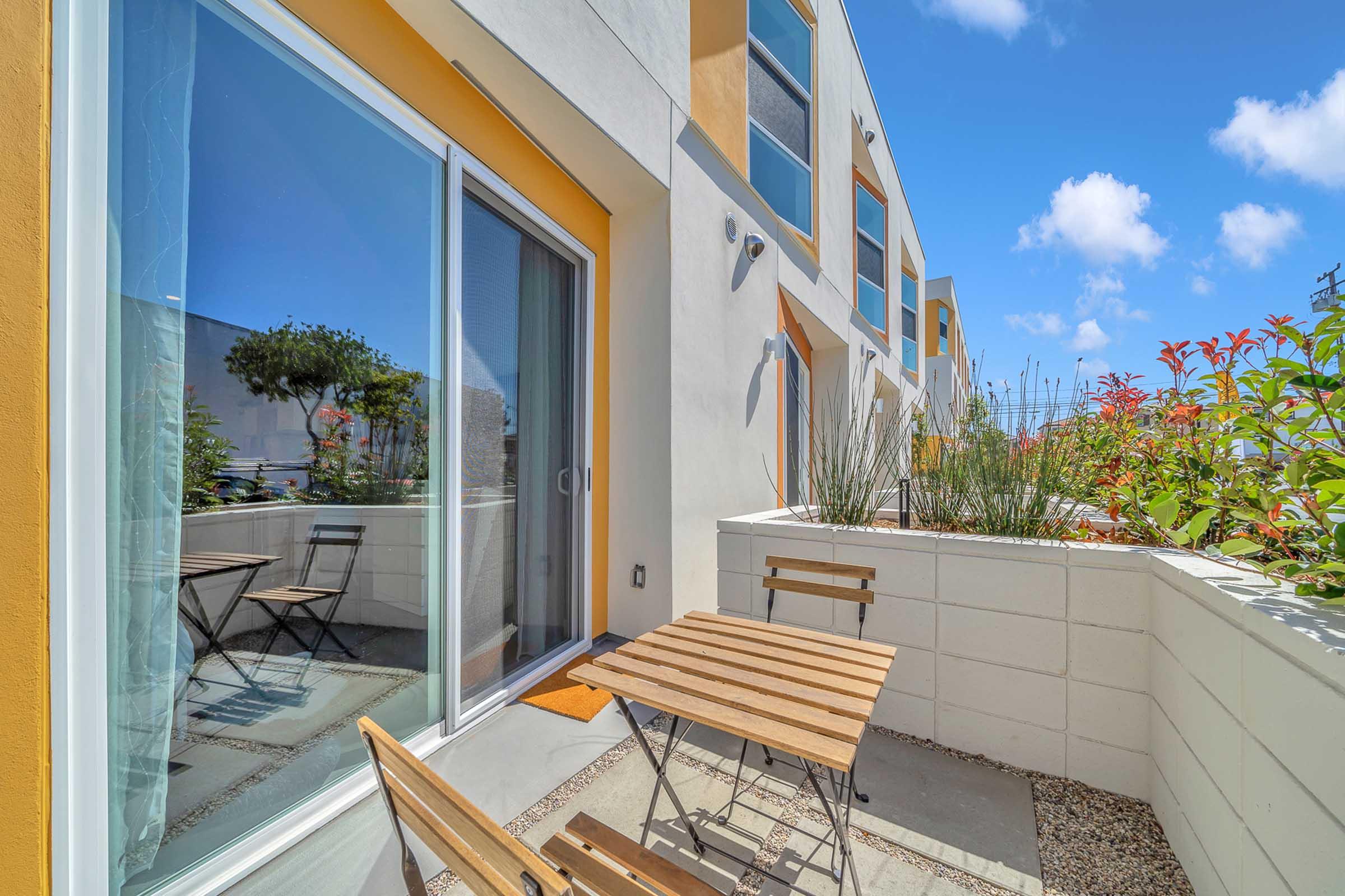 A cozy outdoor patio featuring a wooden table and two chairs, surrounded by colorful plants and modern architecture. The patio is bathed in sunlight, with large sliding glass doors reflecting the sky and landscape. Ideal for relaxation or outdoor dining.