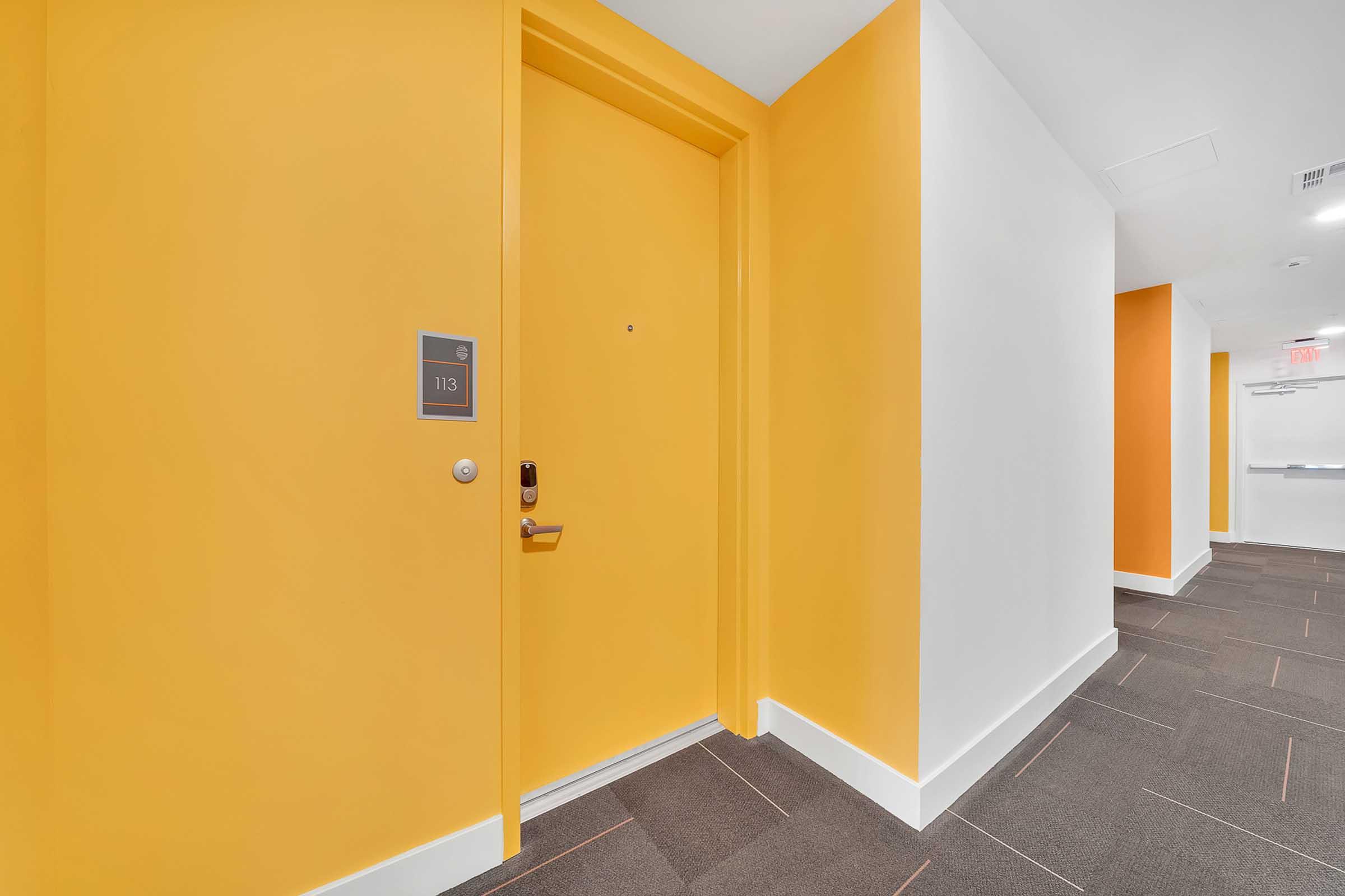 A brightly colored corridor featuring a vibrant yellow wall and an orange accent wall. The focus is on a doorway labeled "113" with a silver handle, situated on the yellow wall. The floor is covered with dark gray carpeting, contrasting with the wall colors, and there is a faint view of the continuation of the corridor beyond.