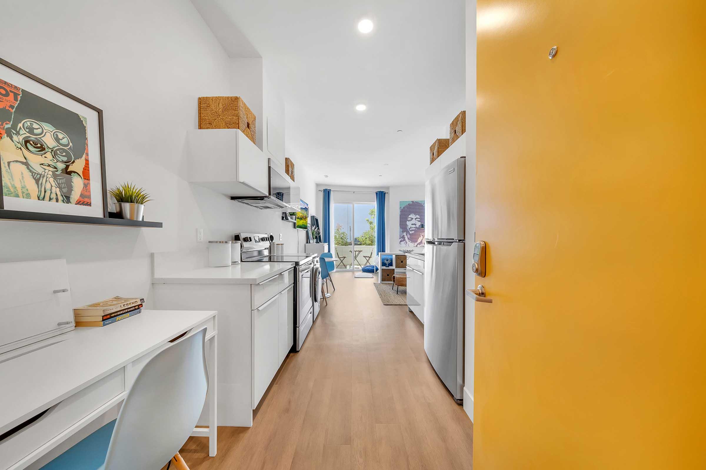 A modern kitchen area with white cabinetry and appliances, featuring a small desk with a blue chair. The hallway leads to a bright living space with large windows, adorned with plants and artwork. The entrance door is bright yellow, adding a pop of color to the interior.