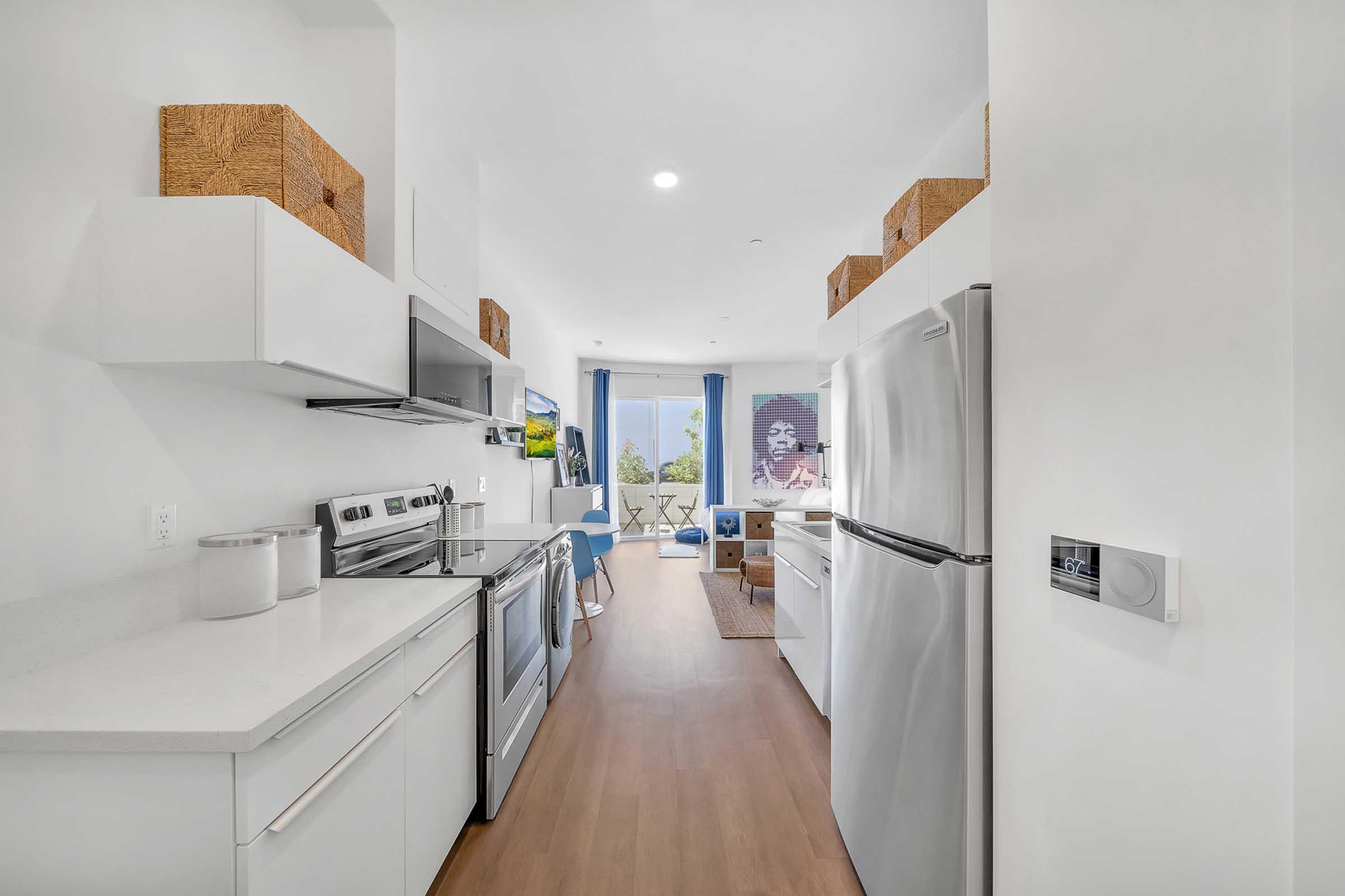 Modern kitchen with stainless steel appliances, including a refrigerator and oven. Brightly lit space features white cabinets, a countertop, and decorative baskets on top. A dining area with a table and chairs is visible in the background, along with a large window allowing natural light.