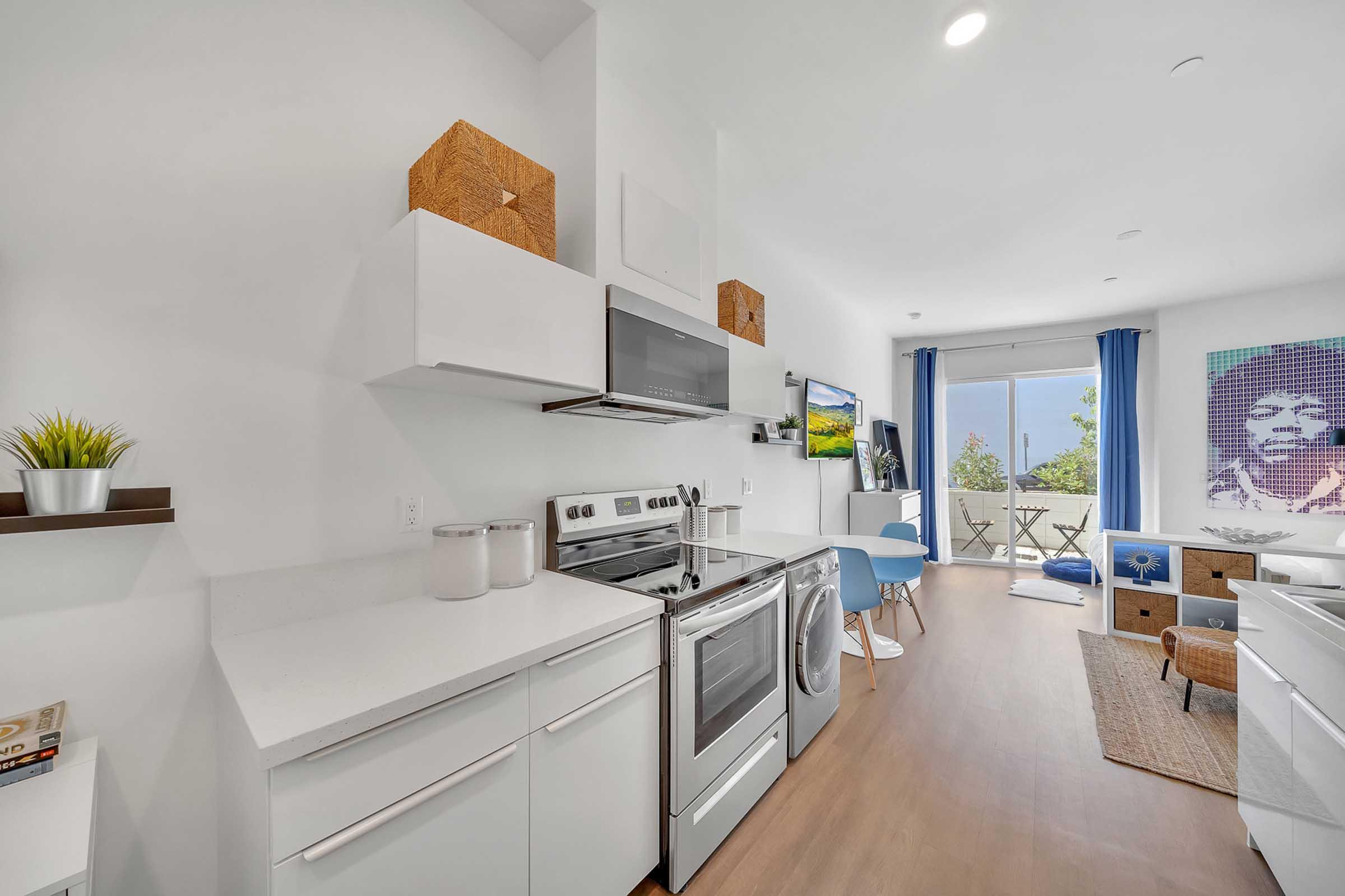 Modern kitchen featuring stainless steel appliances, including a stove, microwave, and washing machine. The space has white cabinetry, minimalistic decor, and a small dining area with blue seating. A large window offers a view of the outdoors, and artwork is displayed on the wall.