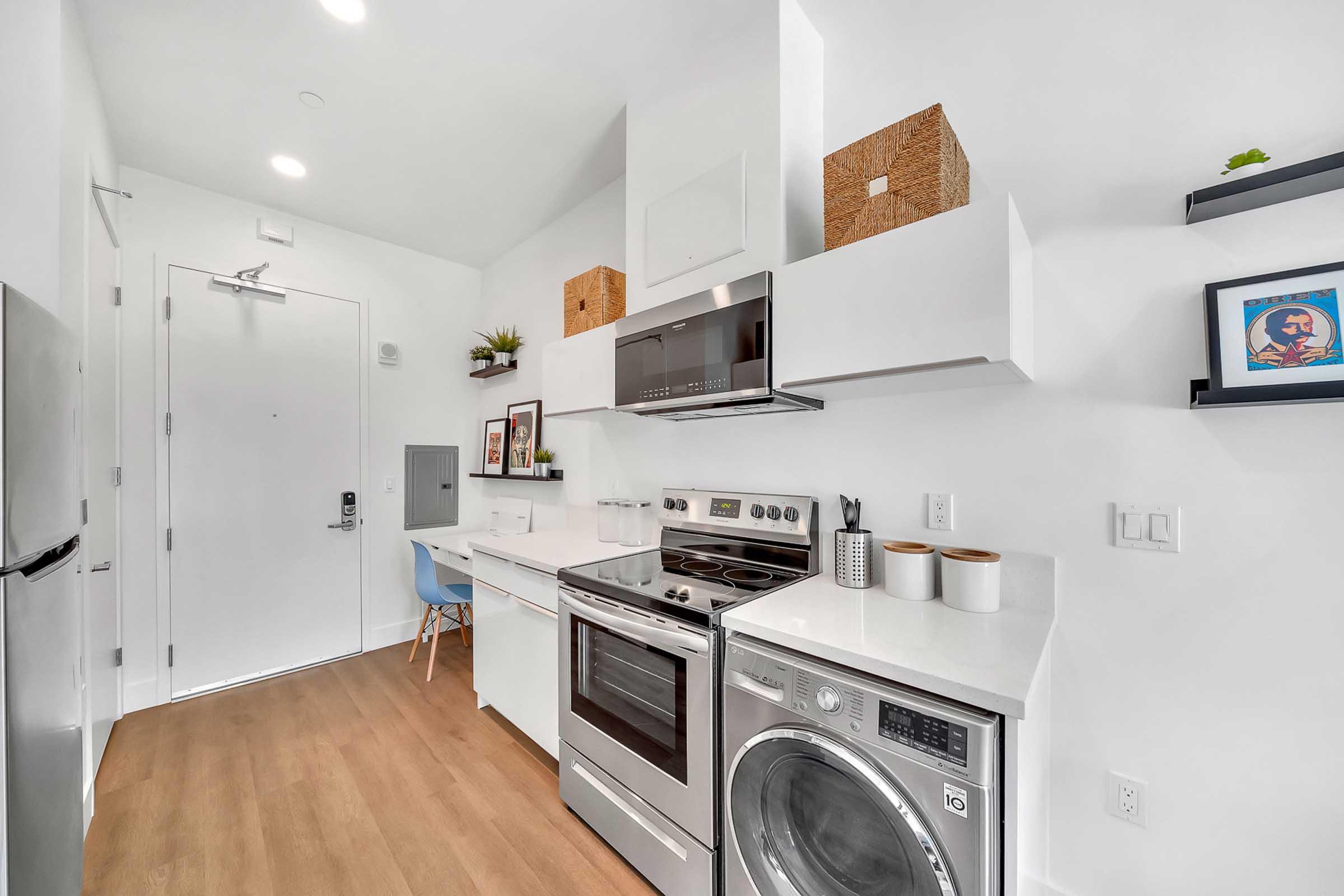 Modern kitchen showcasing a sleek design with white cabinets, an oven, a microwave, a washer/dryer unit, and light wood flooring. The space includes decorative items like baskets and framed pictures, with a small blue chair visible near the entrance. Bright, well-lit environment.
