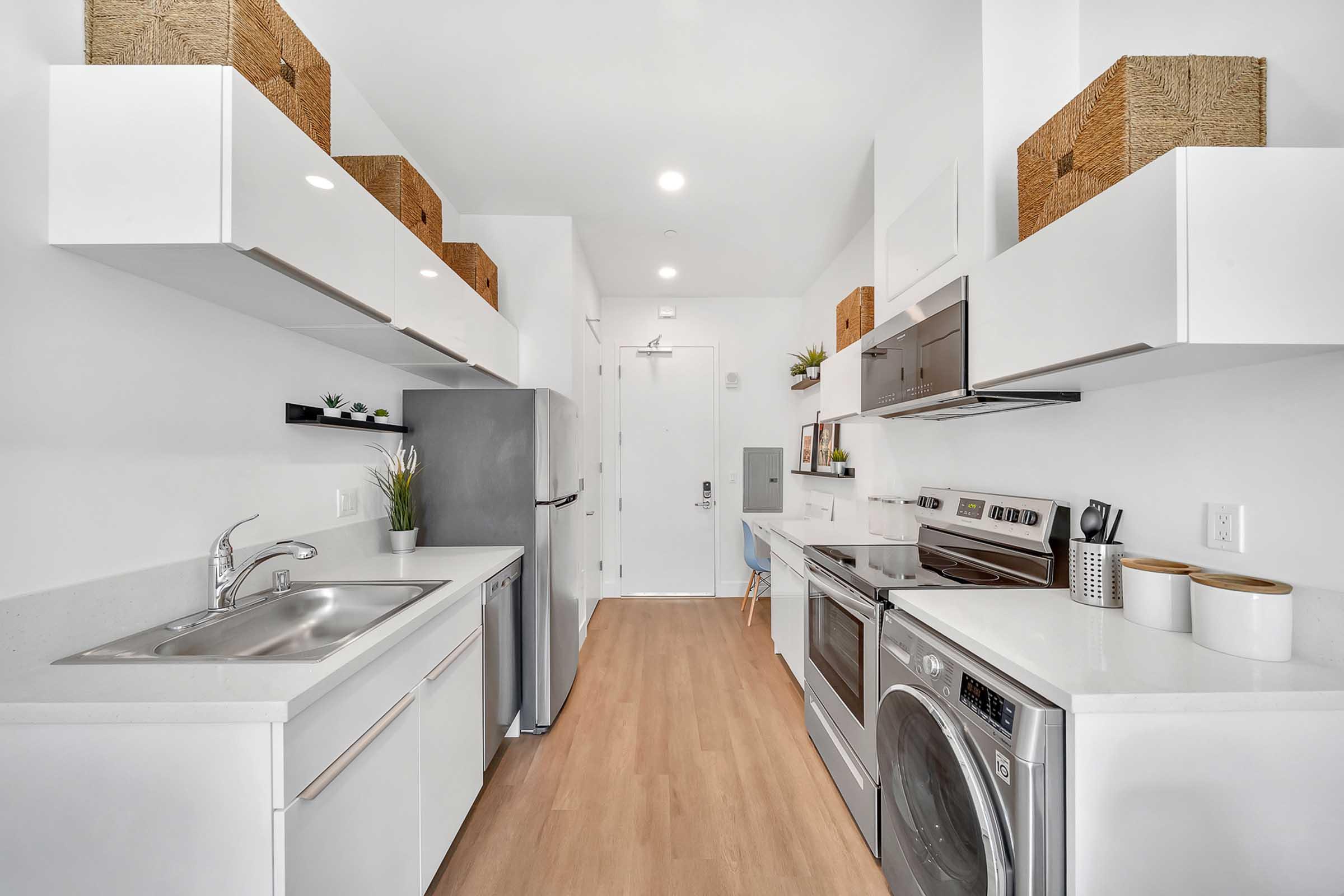 A modern kitchen featuring white cabinetry, stainless steel appliances, and a sleek countertop. The space includes a sink, stove, refrigerator, and a washer/dryer combo. Decorative baskets are placed on top shelves, adding a touch of warmth to the minimalist design. The floor is light wood, enhancing the airy feel.