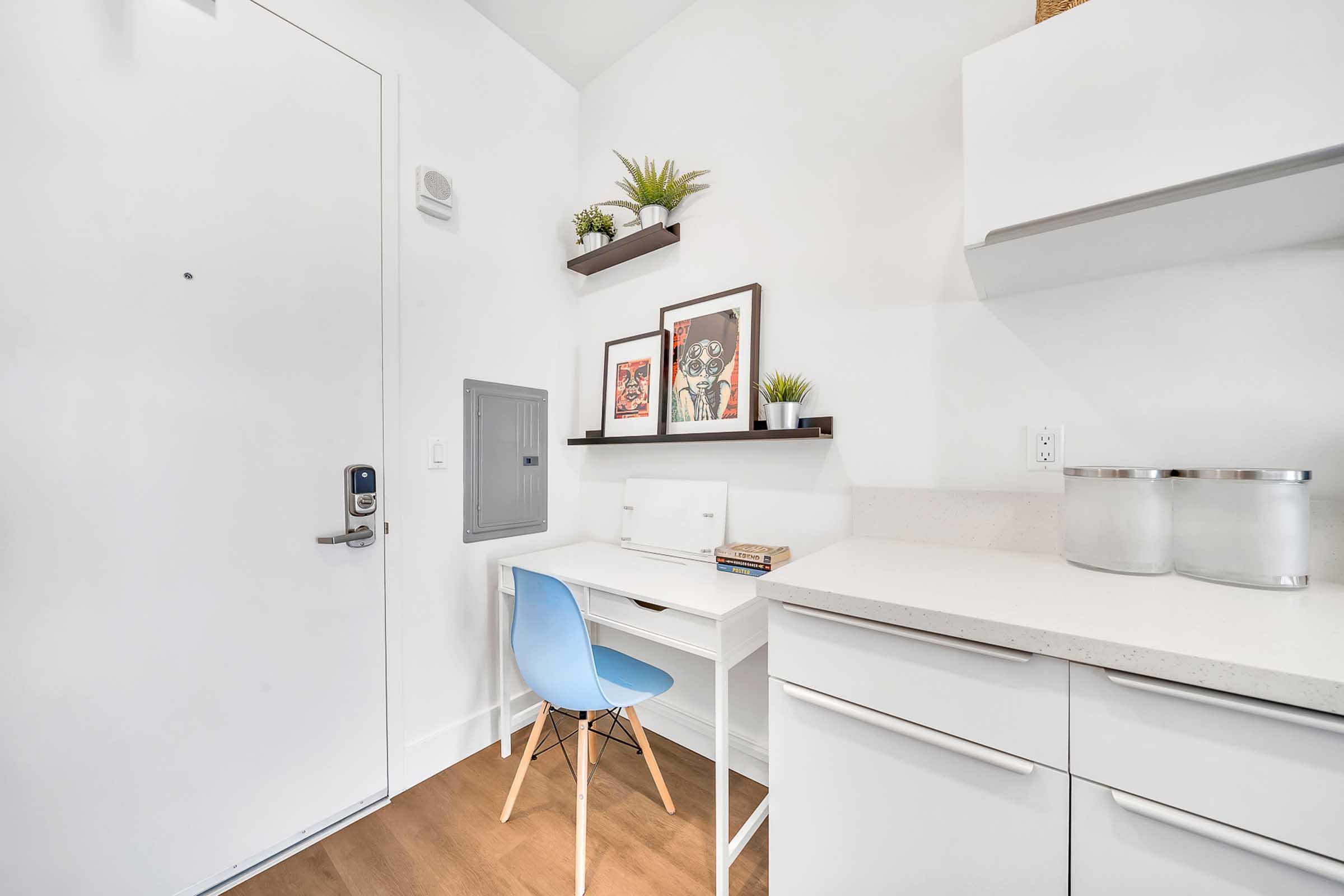 A small home office space features a sleek white desk with a blue chair, a mounted shelf with decorative plants, and framed artwork on the wall. A white cupboard with drawers is visible, alongside a wall-mounted mailbox. The overall aesthetic is modern and minimalist, with light wood flooring and bright walls.