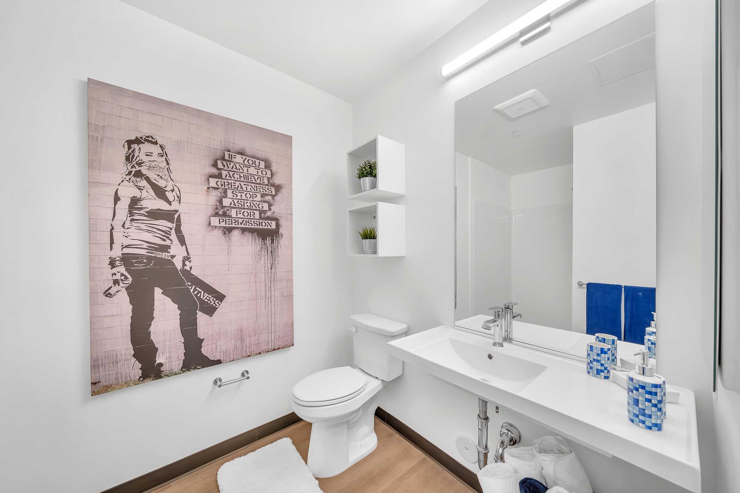 A modern bathroom featuring a wall-mounted artwork depicting a figure with text. The decor includes a clean white sink, a toilet, and minimalist shelving with small potted plants. The floor is light wood, and there are blue and white accessories in the space, creating a fresh and contemporary atmosphere.
