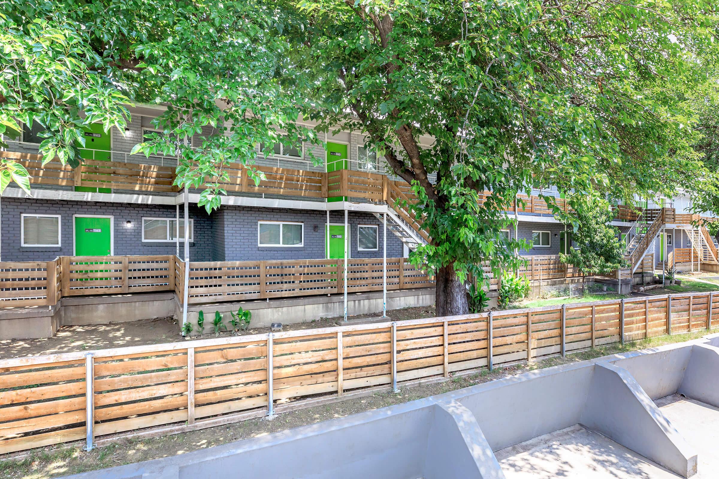 A row of multi-unit residential buildings with green doors, surrounded by trees. The front area features wooden fencing and a landscaped yard, providing a spacious and inviting atmosphere. The buildings have outdoor staircases, offering access to the upper levels. The scene is bright and well-maintained.