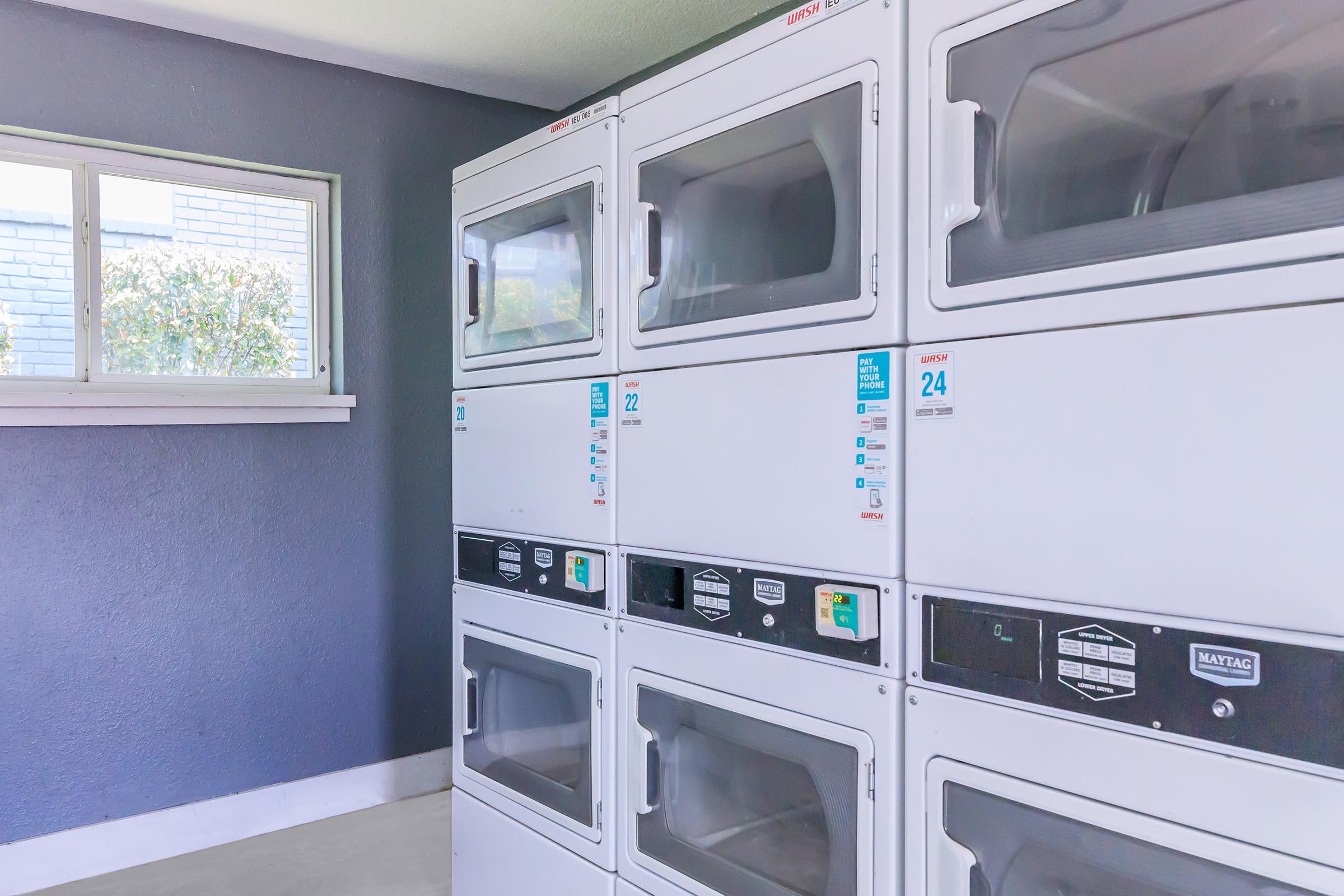 A brightly lit laundry room featuring stacked white washers and dryers with digital controls. The wall is painted a dark blue, and there’s a window allowing natural light to enter. The appliances are neatly arranged, showcasing their clean and modern design.