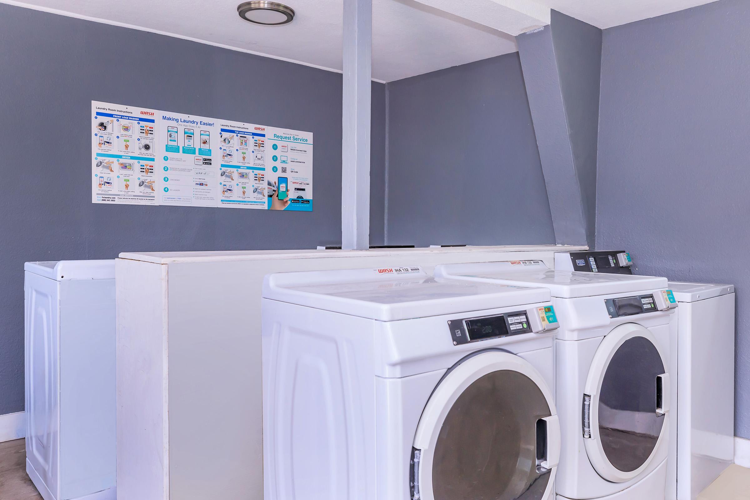 A laundry room featuring several white washing machines and dryers. A poster on the wall provides instructions for using the laundry facilities. The room has gray walls and a ceiling light, with a clean and organized appearance.