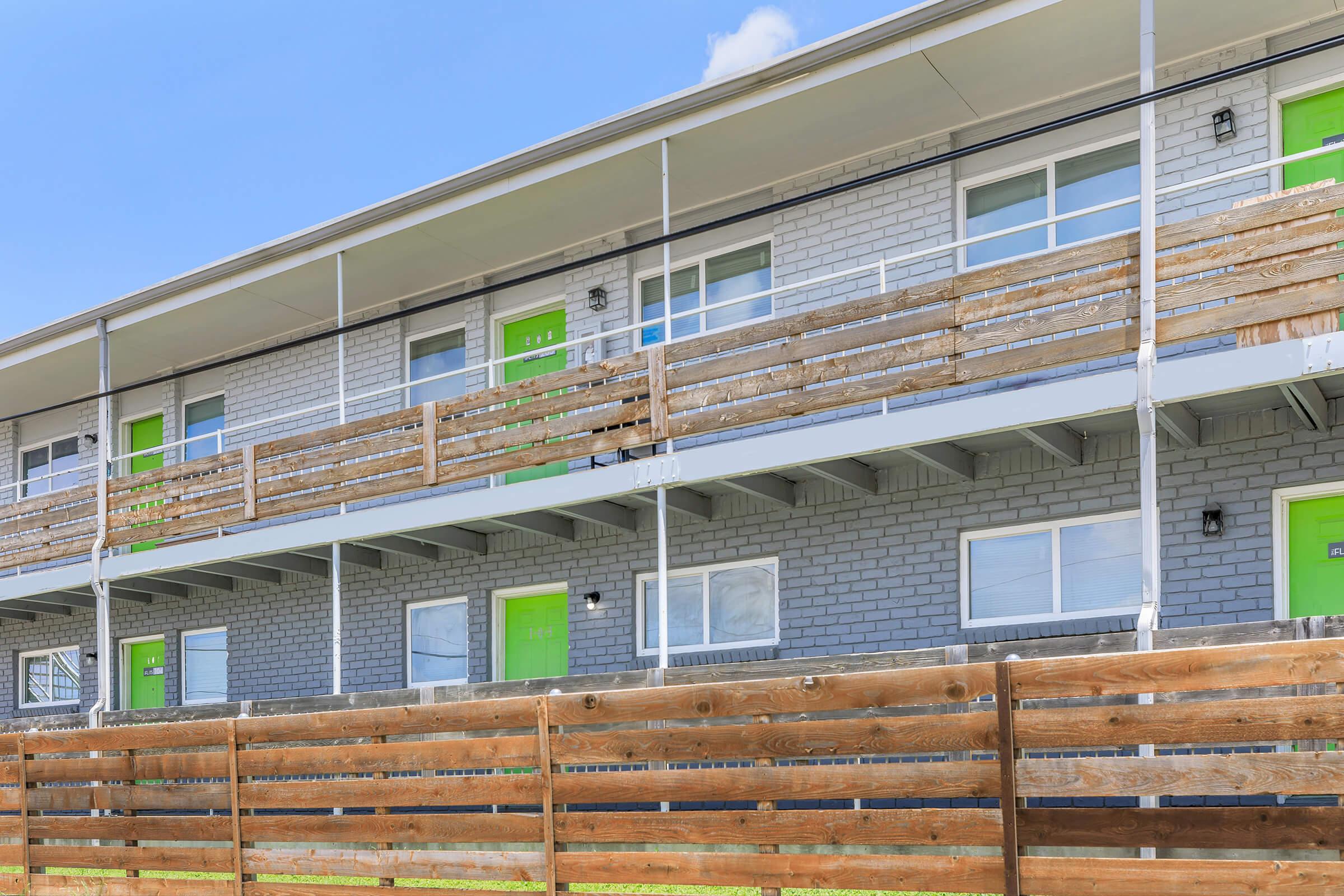 Two-story apartment building with gray brick walls and bright green doors. Each unit has a small balcony with wooden railings. A wooden fence is visible in the foreground, separating the building from a grassy area. The sky is clear and blue, adding a vibrant atmosphere to the scene.